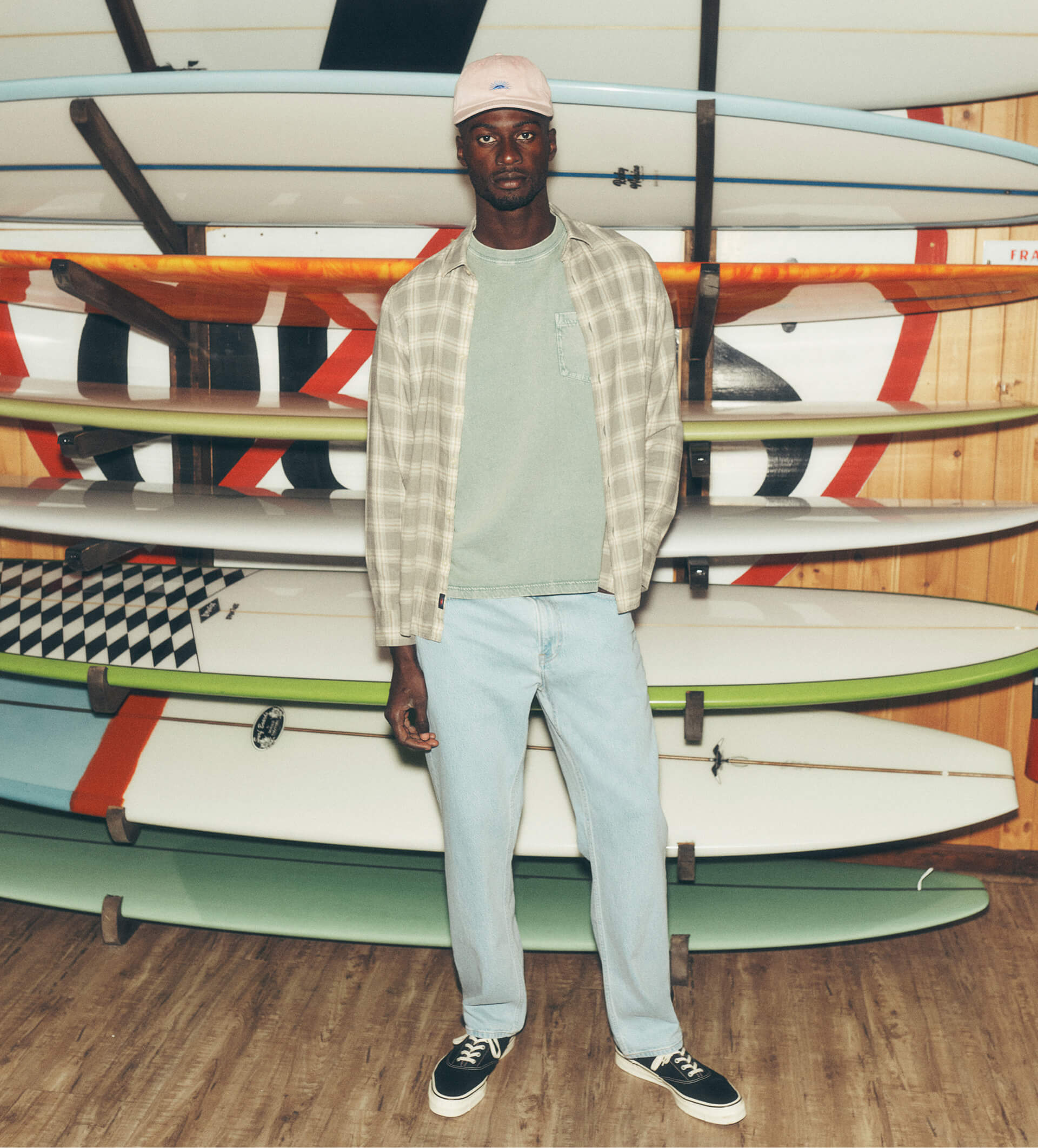 A male model wearing stretch terry pants, a dad cap, an open plaid button down shirt  over a sunwashed tee and vans sneakers standing in front of a rack of surf boards.