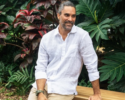 Man sitting on a bench in front of foliage, wearing the Laguna Linen shirt.