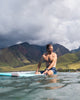 Man sitting on a surfboard in the ocean, wearing shorelite shorts. Mountains in the background.