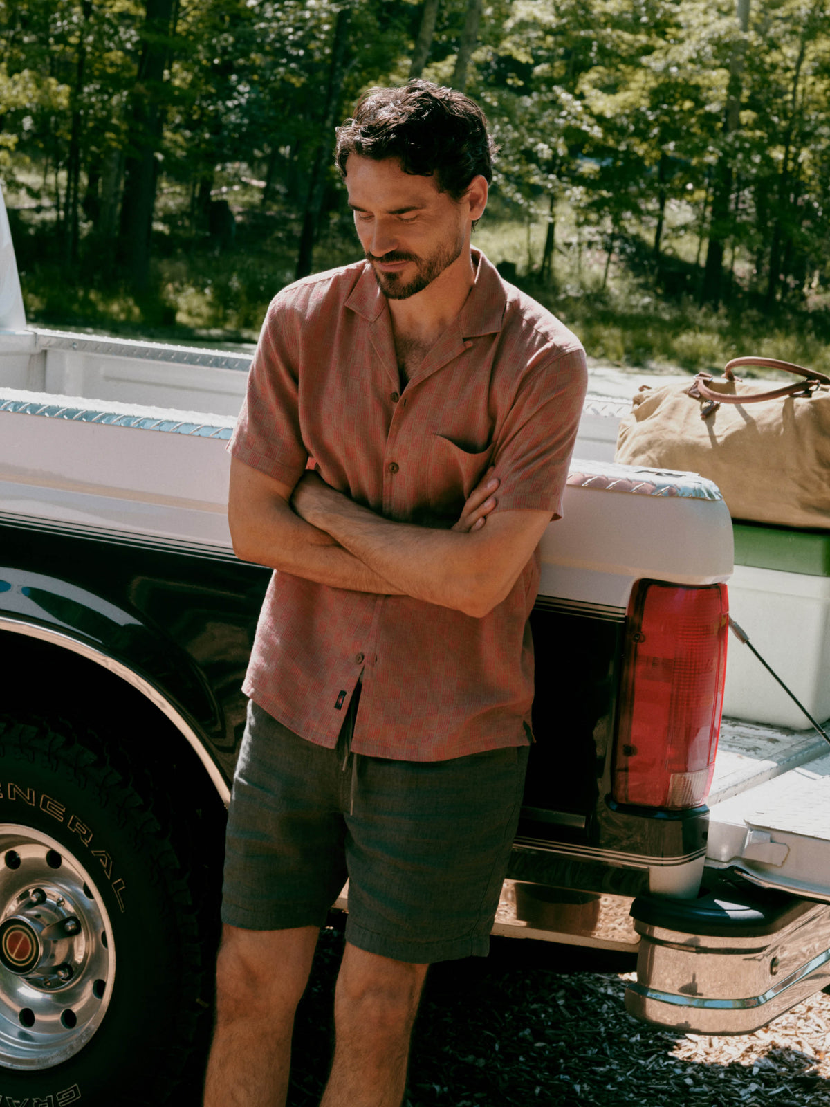 A man stands leaning against the back of a white pickup truck outdoors, wearing a muted dusty rose short-sleeve button-up shirt and dark olive-gray shorts. Trees and gravel are visible in the background, along with a tan duffel bag in the truck bed., image 1 of 7