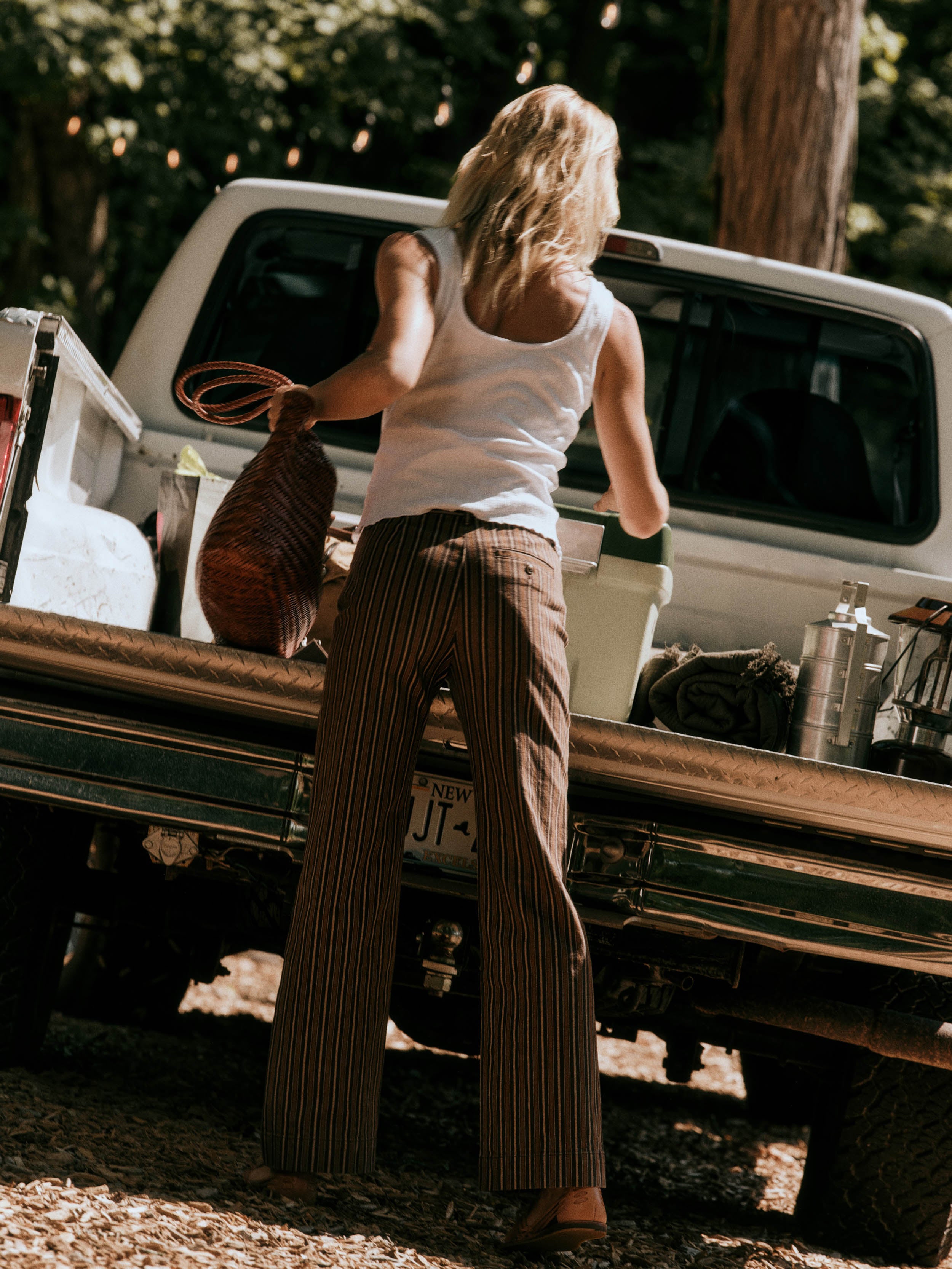 A woman stands with her back to the camera, loading or unloading items from the bed of a white pickup truck in a wooded outdoor setting. She is wearing a white tank top and brown pinstriped wide-leg pants, holding a brown woven bag.