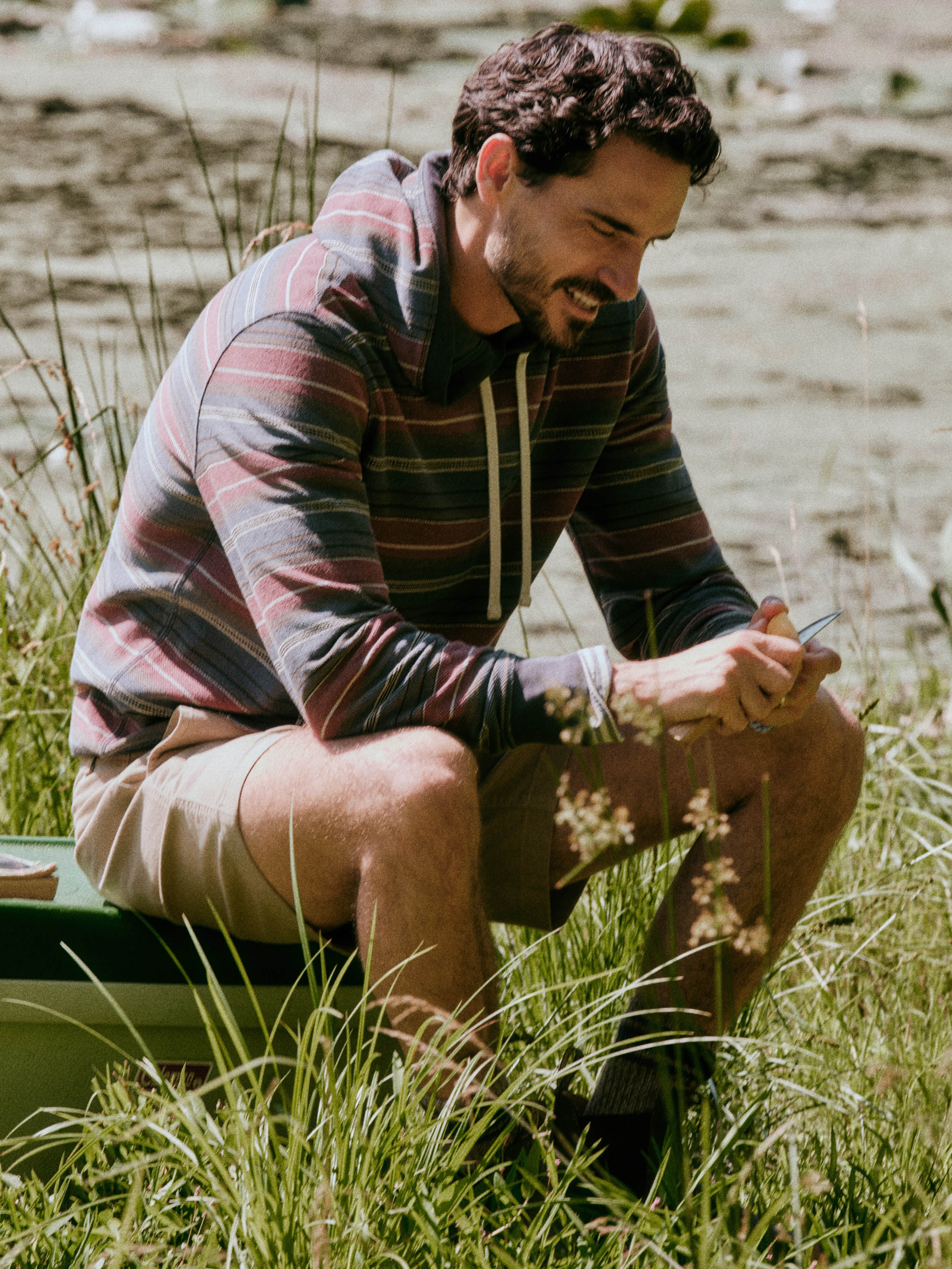 A man wearing a striped long-sleeve hoodie and beige shorts sits in tall grass outdoors, looking down at a phone.