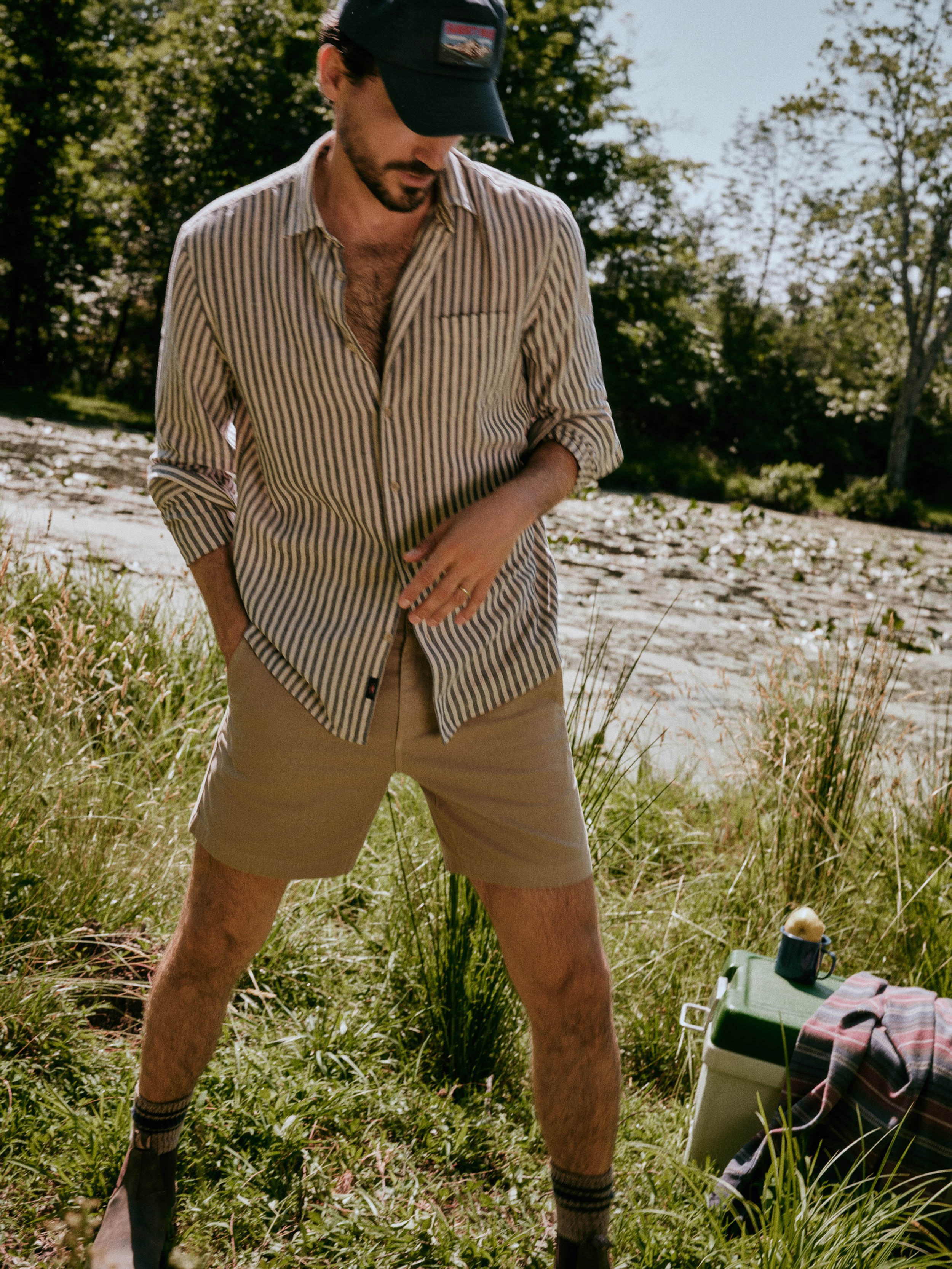 A man wearing a blue and white vertically striped button-down shirt, tan shorts, and a black cap stands outdoors near a river with a cooler and blanket on the grass beside him.
