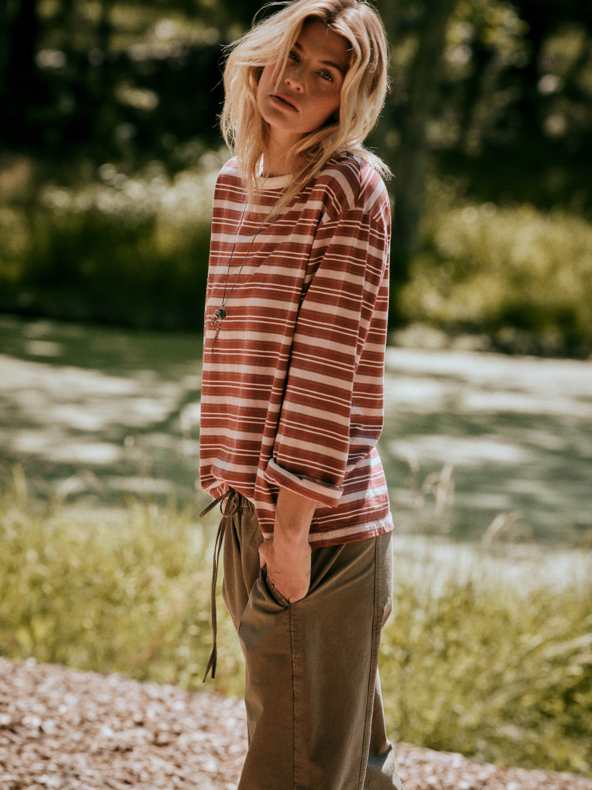 A woman standing outdoors in a wooded area, wearing a red and light pink striped long-sleeve top with a drawstring hem and olive green wide-leg pants.