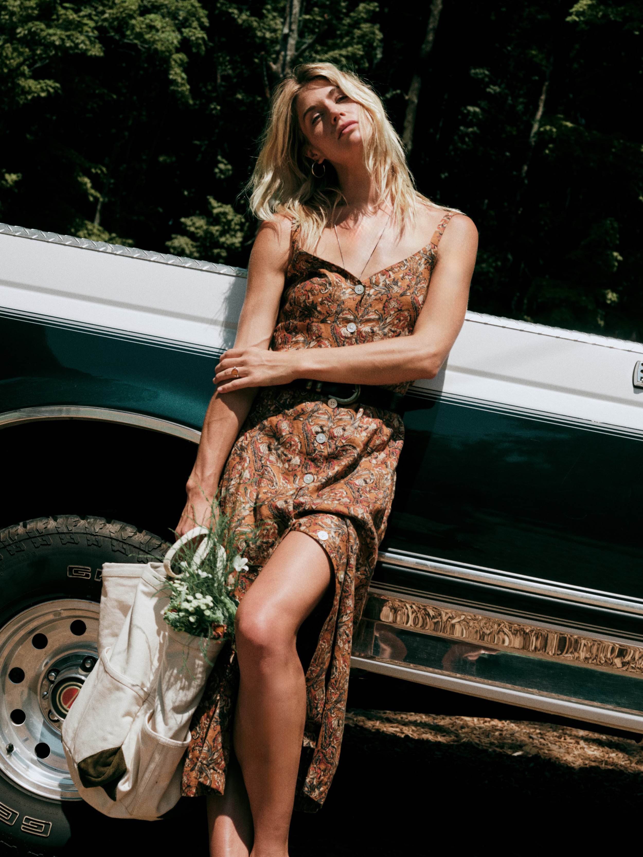 A woman wearing an orange floral print spaghetti-strap dress leans against the side of a vintage teal and white pickup truck outdoors, holding a cream tote bag with flowers.