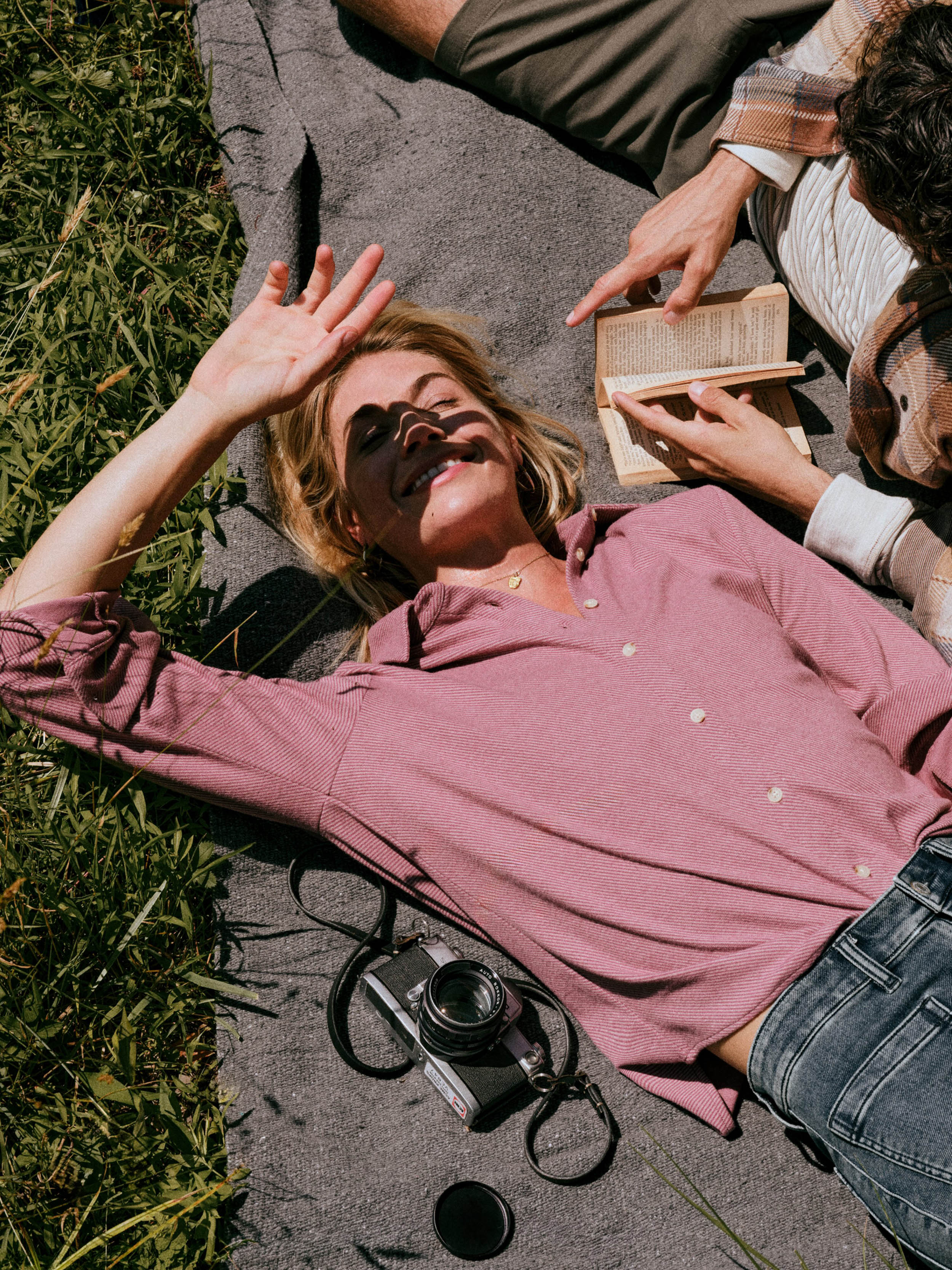 A woman lying on a gray blanket outdoors on grass, wearing a dusty rose button-up shirt and blue jeans, with another person nearby holding an open book. A vintage camera and a small black accessory rest on the blanket.