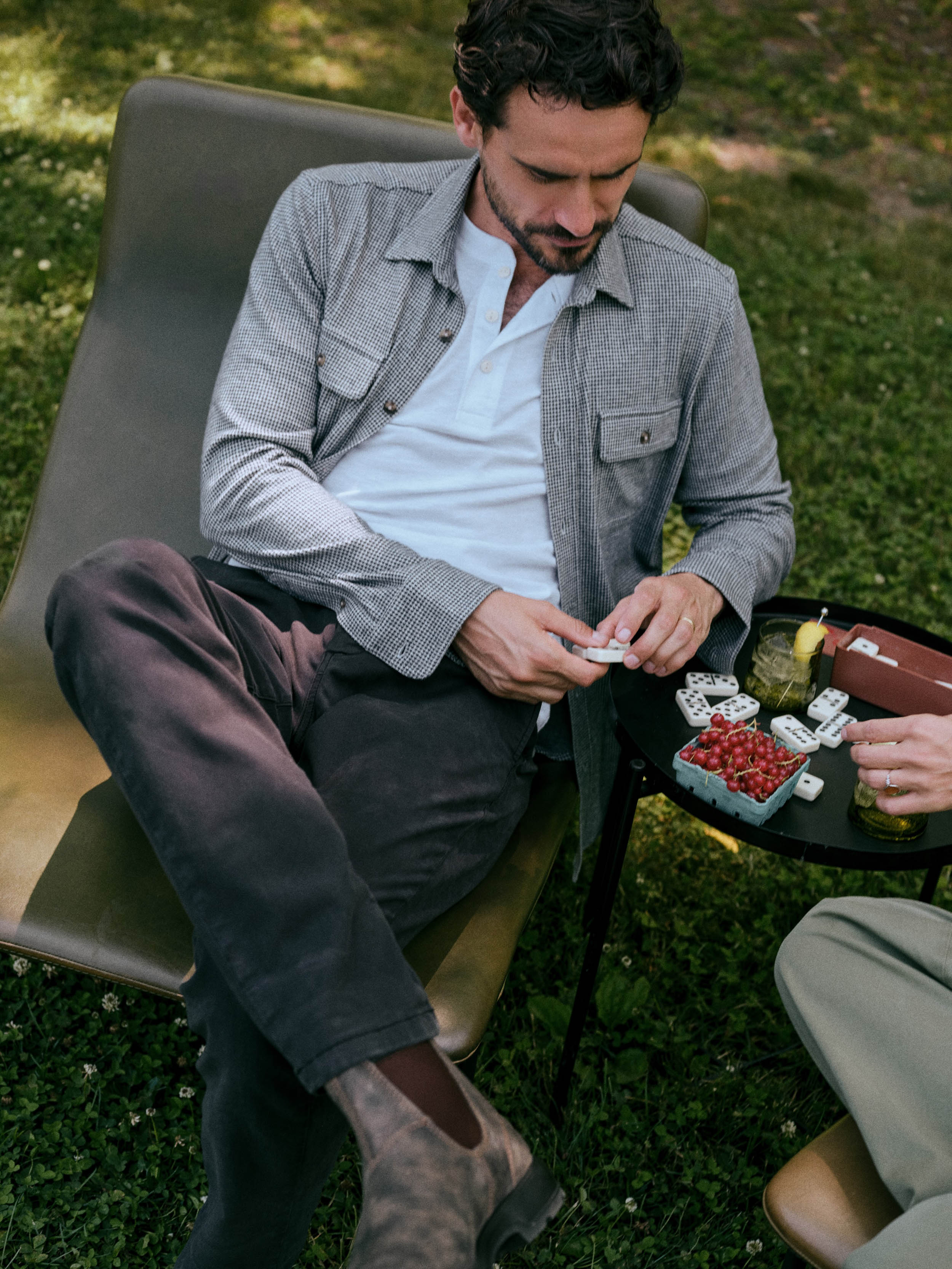 A man sits in a lounge chair outdoors on grass, wearing a light gray overshirt over a white polo shirt and dark trousers. A small table beside him holds cards and a drink.