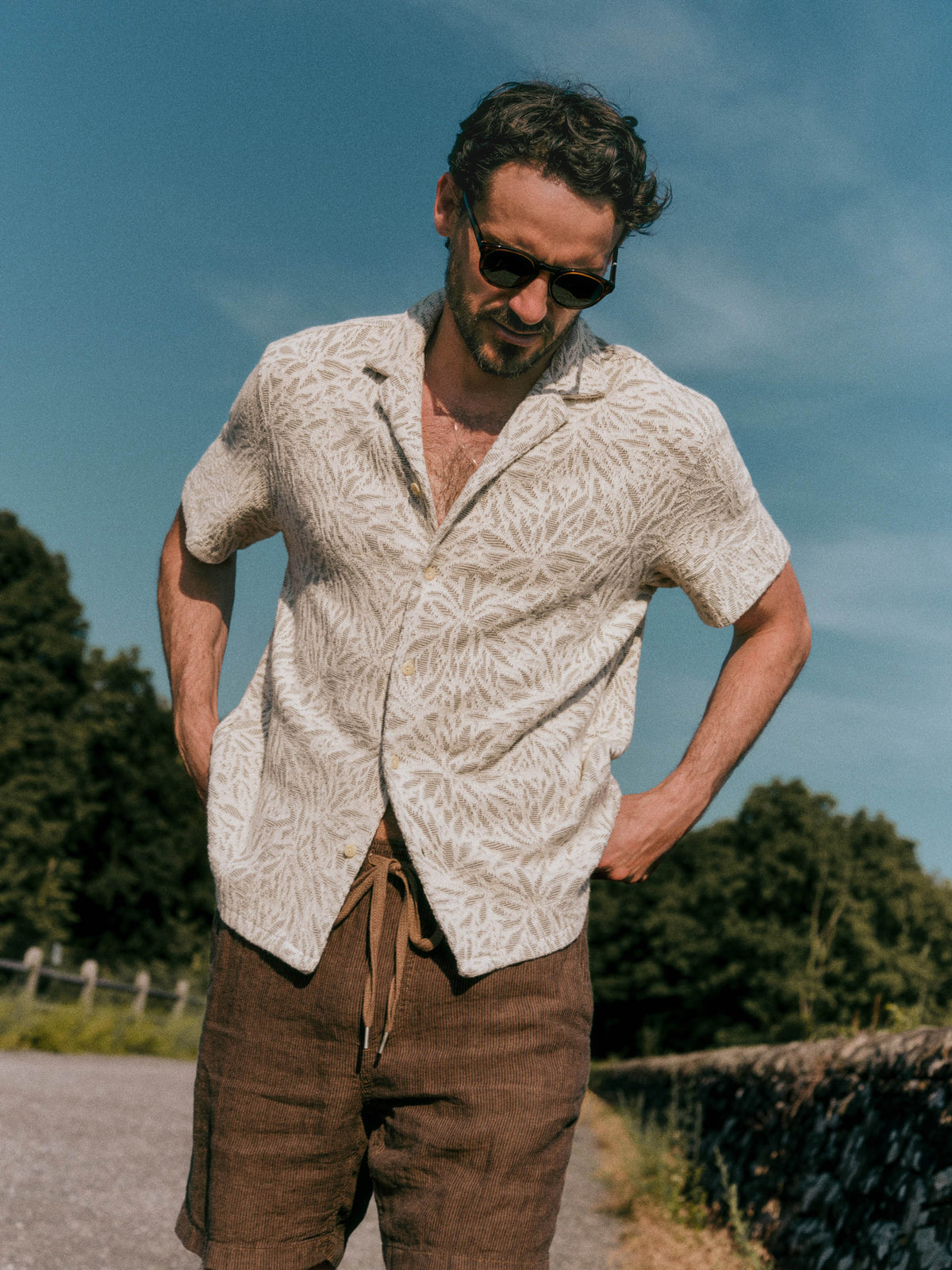 A man wearing a short-sleeve button-up shirt with a light green and white leaf pattern, paired with brown pants, standing outdoors on a sunny day with trees and a blue sky in the background., image 1 of 7