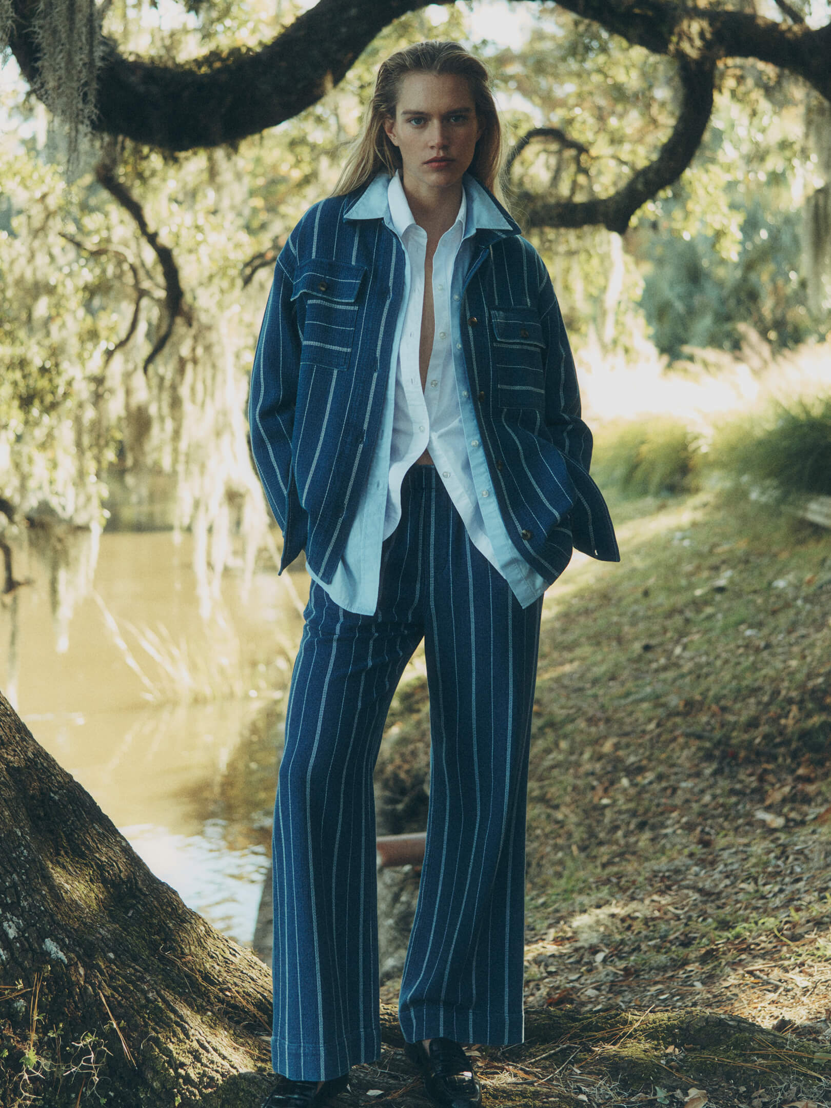 A female model in a Charleston garden, surrounded by spanish moss and a pond, wearing an indigo striped jacket and matching bottoms, layered over a chambray shirt and a white shirt.