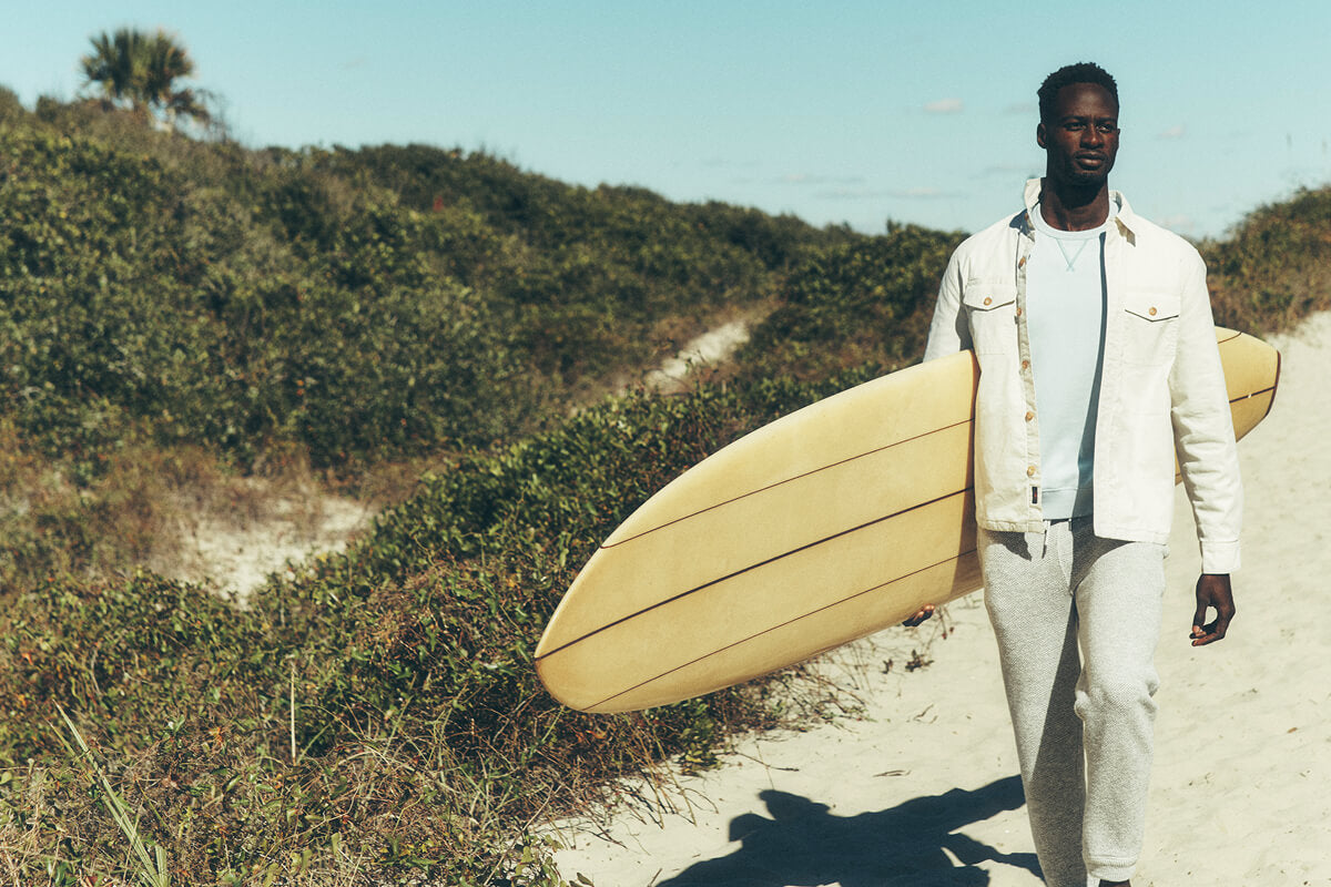 Model wearing surf revival collection carrying a surf board on a path towards the beach