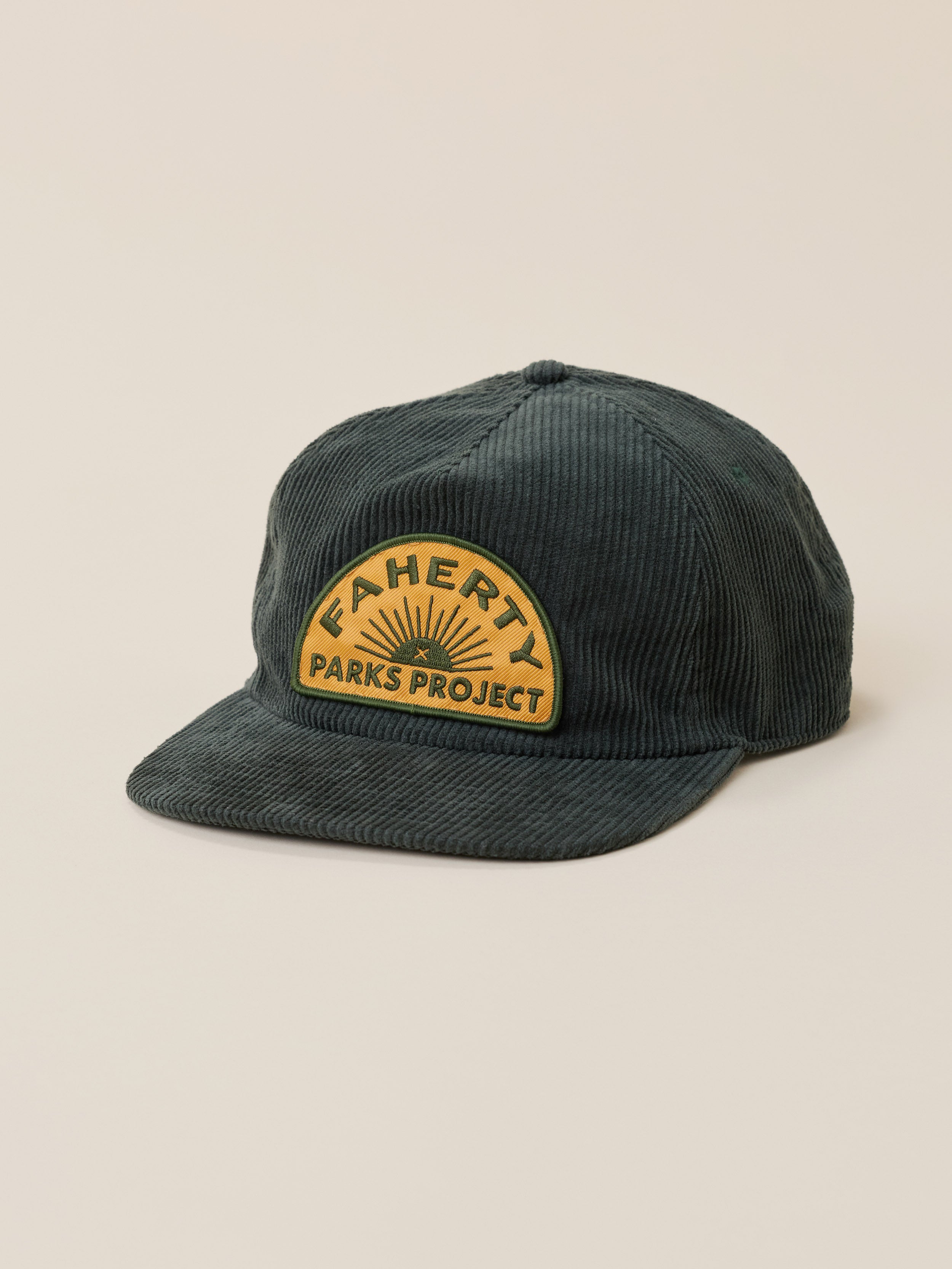 A dark green corduroy baseball cap with a curved yellow patch on the front that reads 'FAHERTY x PARKS PROJECT' with a sunburst design, set against a solid beige background.