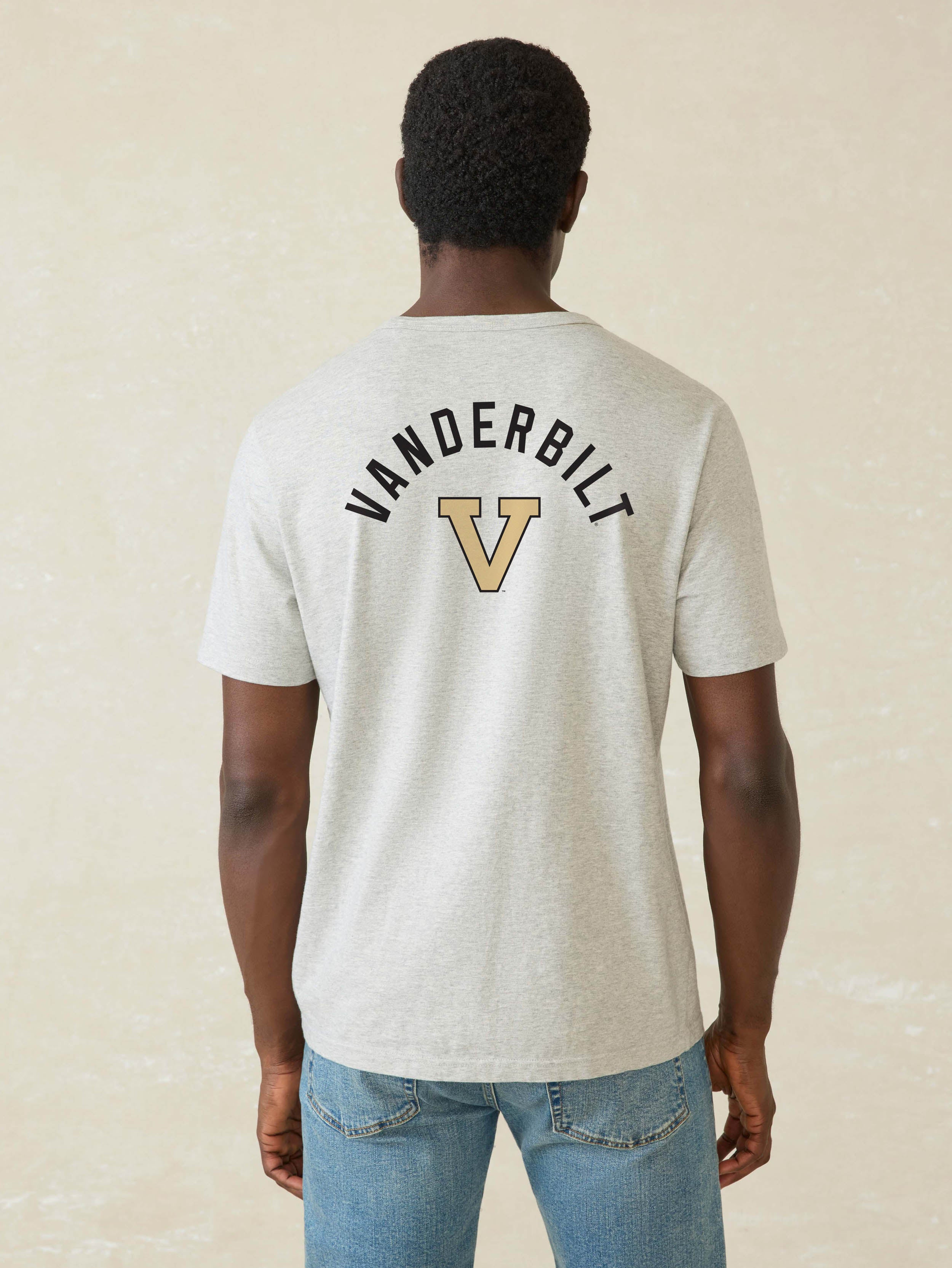 A man viewed from behind wearing a light heather gray t-shirt with a black and gold Vanderbilt logo on the back, paired with blue jeans, against a textured beige background.
