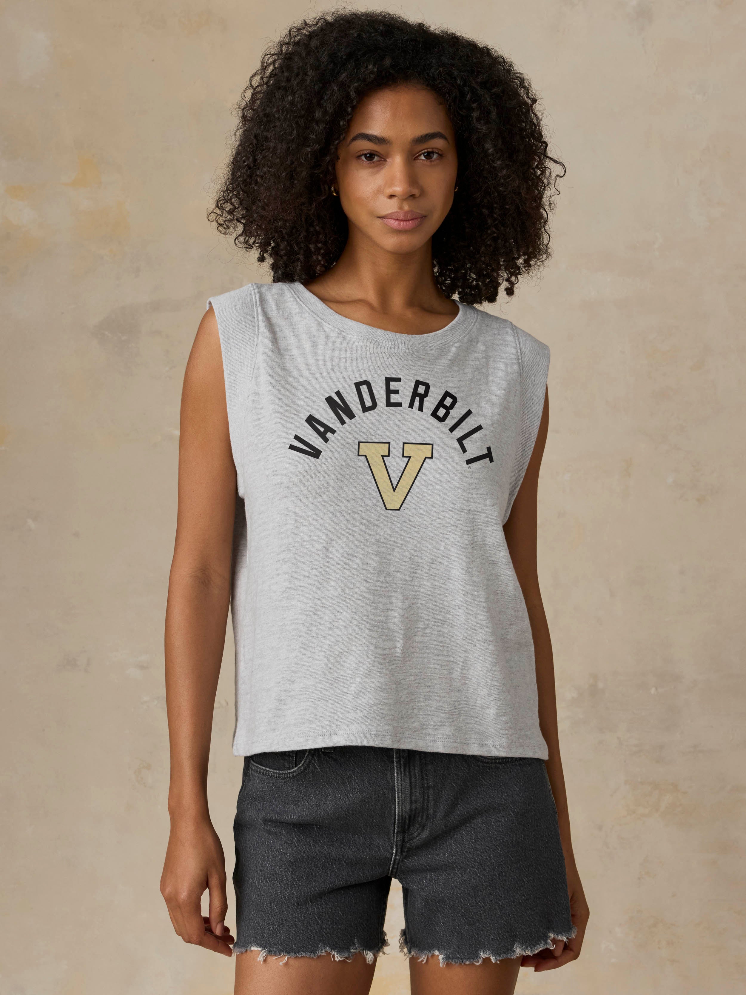 A woman modeling a light gray sleeveless crop top with 'VANDERBILT' and a large 'V' logo printed on the front, paired with dark denim shorts against a textured beige background.