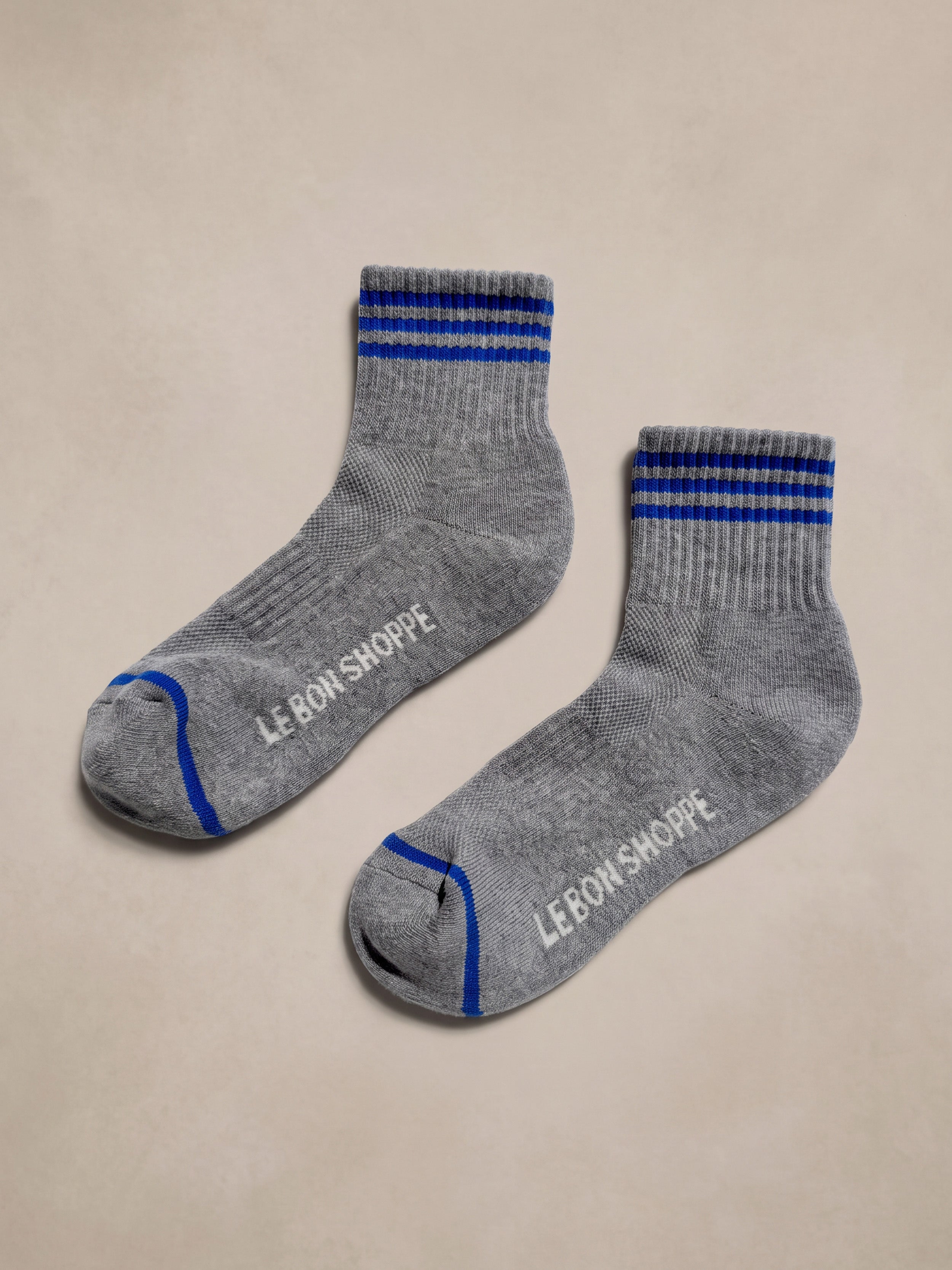 A pair of heather gray ankle socks with blue stripes on the cuff and toe, featuring the brand name 'Le Bon Shoppe' printed in white on the sole, displayed on a neutral beige background.