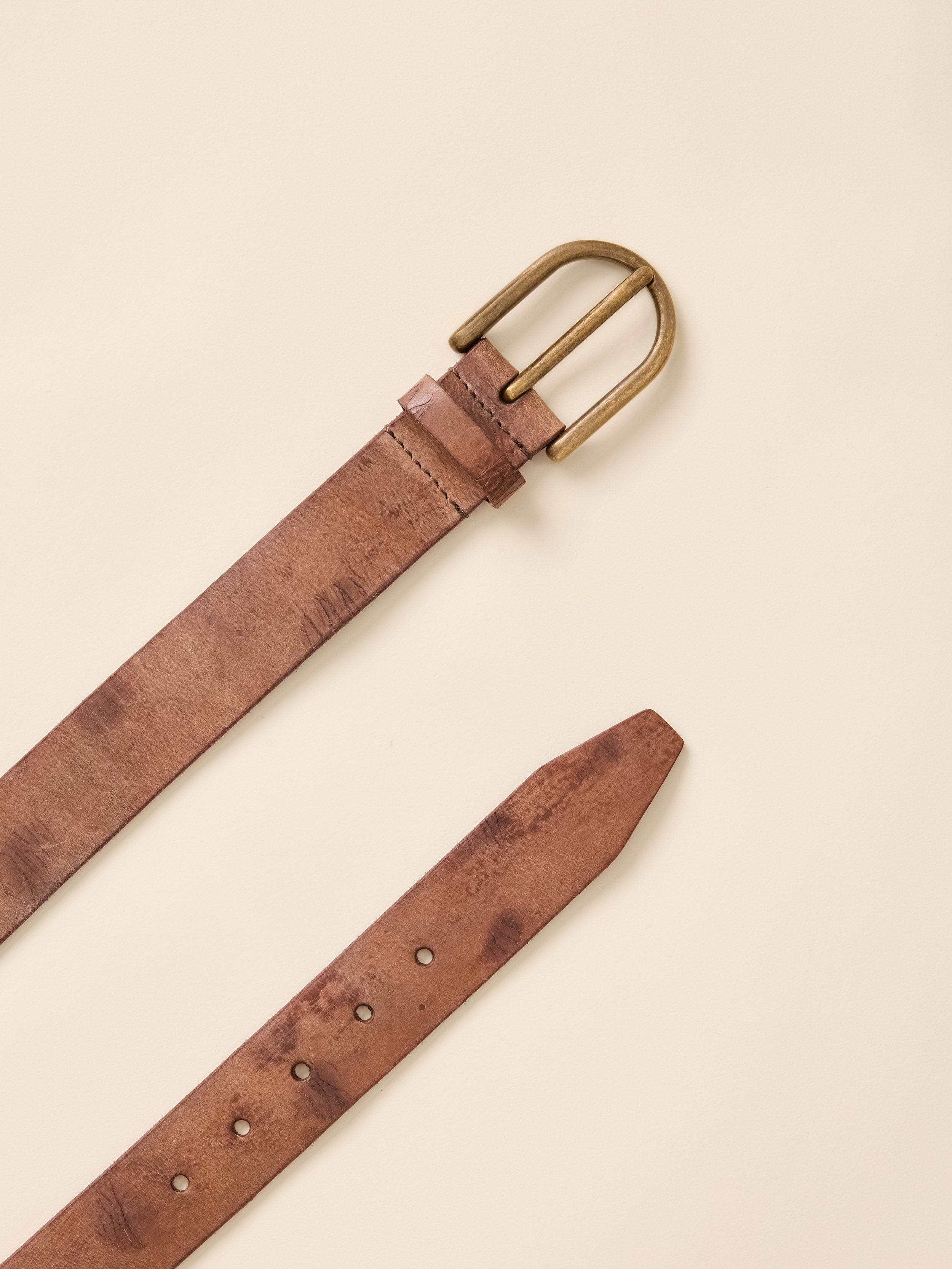 A close-up of a distressed brown leather belt strap with a brass buckle, laid diagonally on a light cream background.