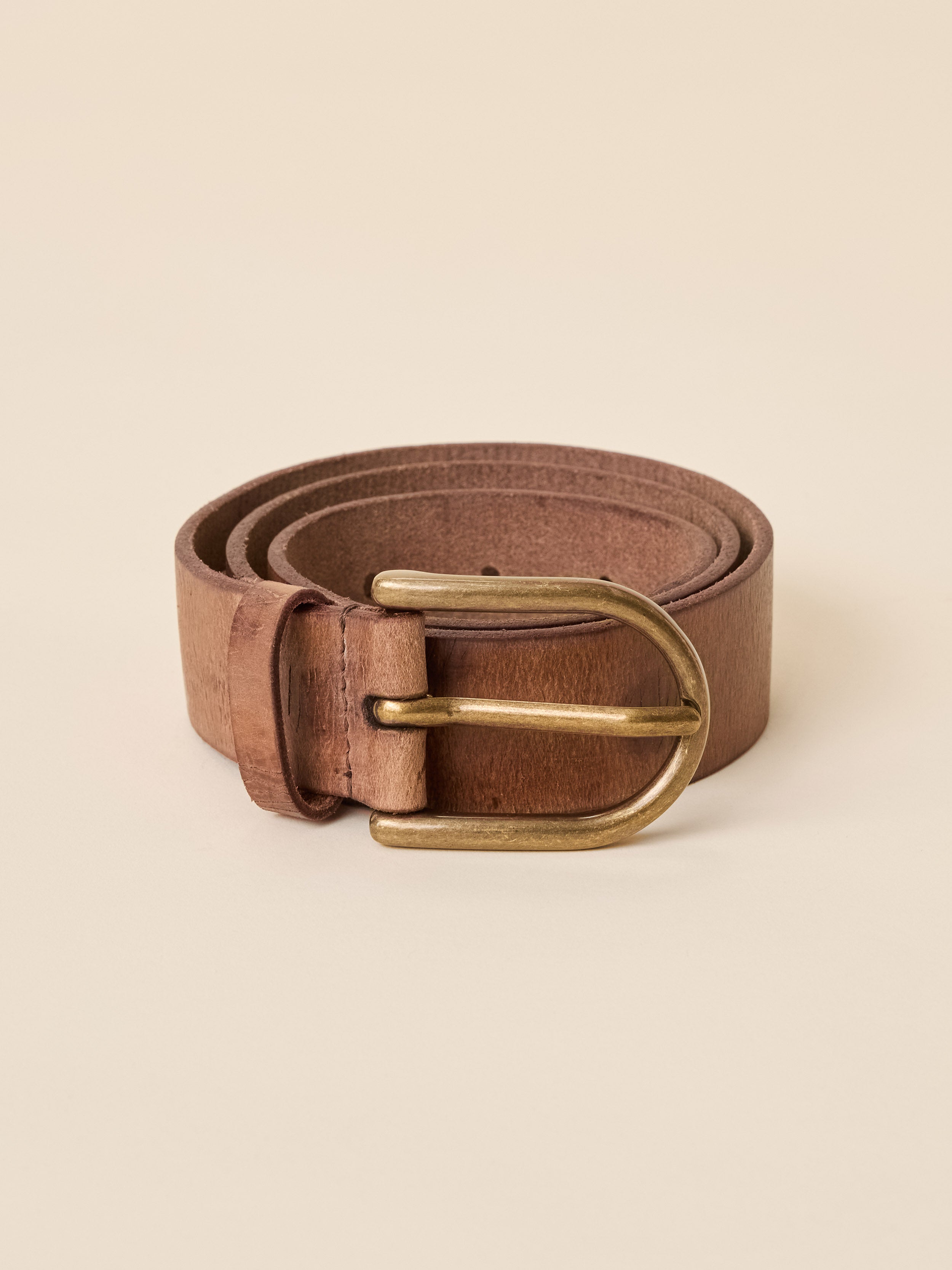 A brown leather belt with a brass buckle, neatly coiled on a solid beige background.