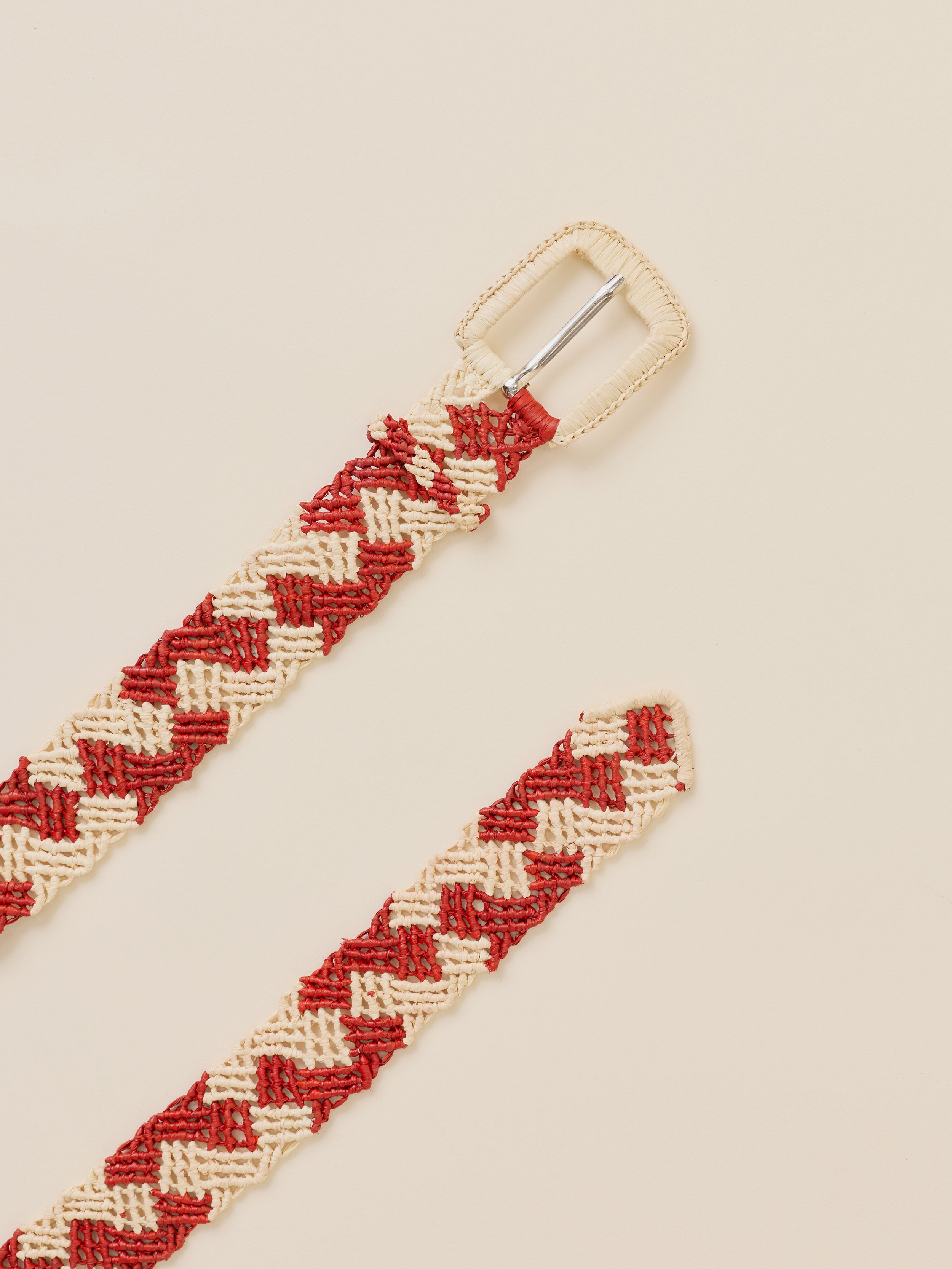 A woven braided belt with a red and cream chevron pattern and a silver metal buckle, shown on a solid light beige background.