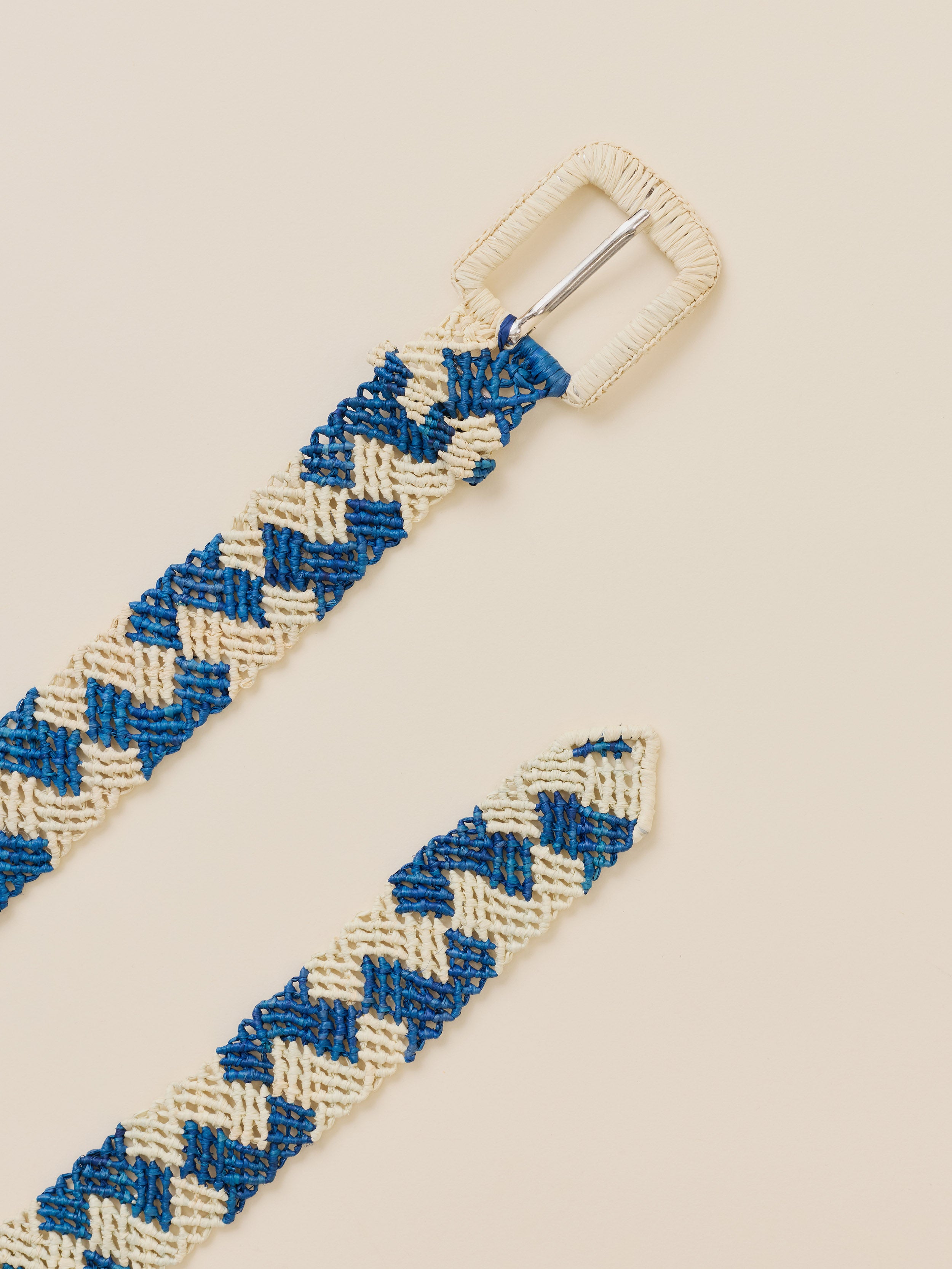 A close-up of a woven belt strap with a blue and cream chevron pattern, featuring a cream-colored metal buckle and a matching woven tip, set against a solid light beige background.
