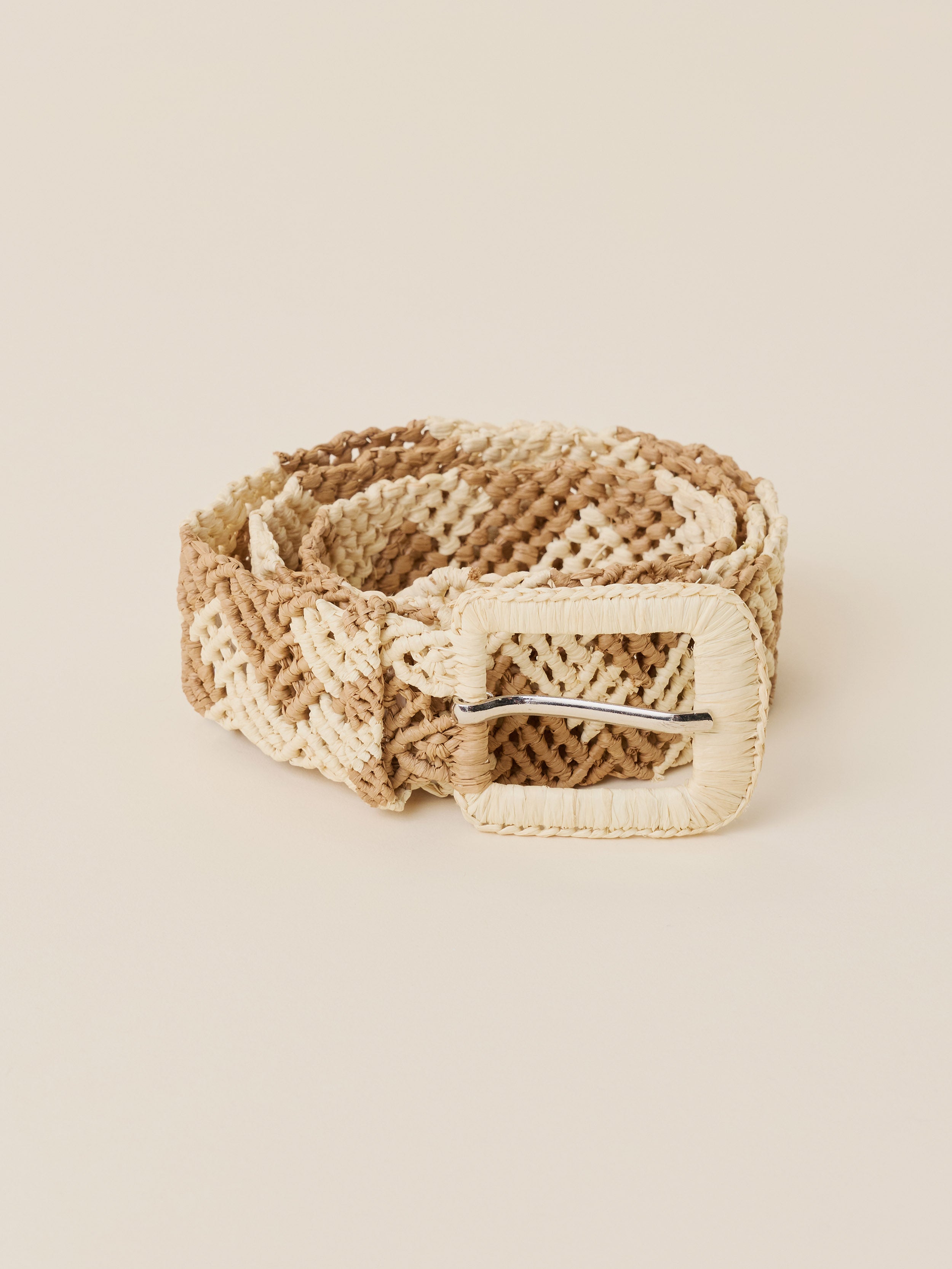 A woven belt with a textured geometric pattern in beige and cream, featuring a large cream-colored buckle with a silver metal bar, set against a solid light beige background.