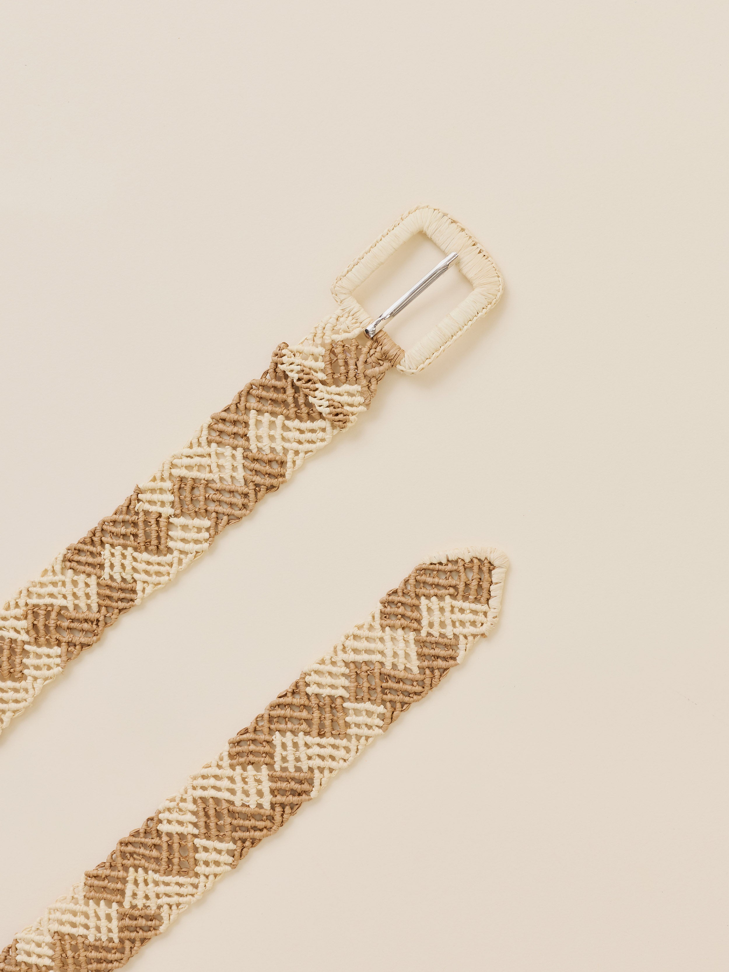 A woven belt with a textured herringbone pattern in beige and brown, featuring a cream-colored rectangular buckle, laid out diagonally on a solid cream background.