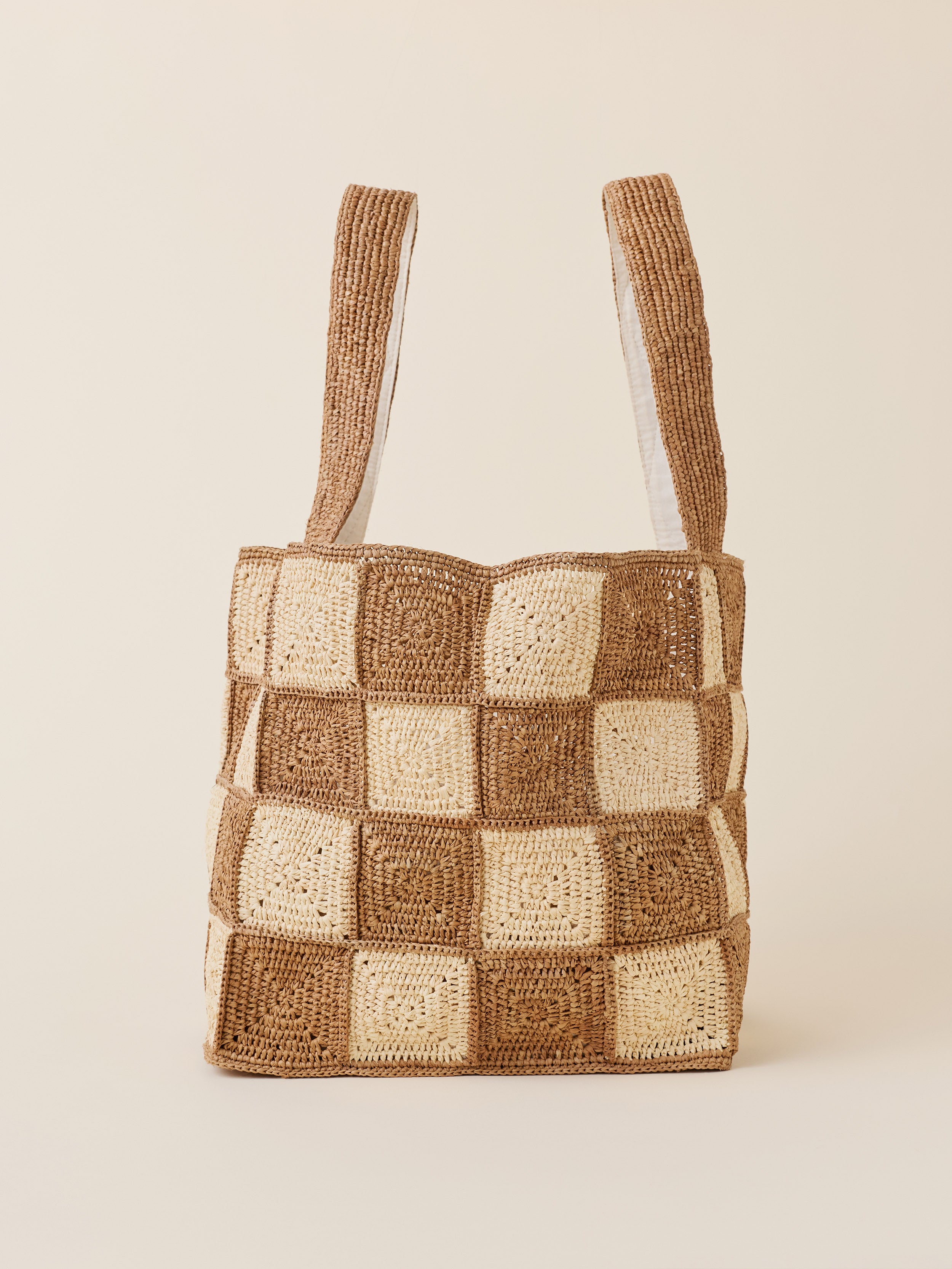 A raffia tote bag with a checkerboard pattern in brown and beige, featuring two shoulder straps, set against a solid light beige background.