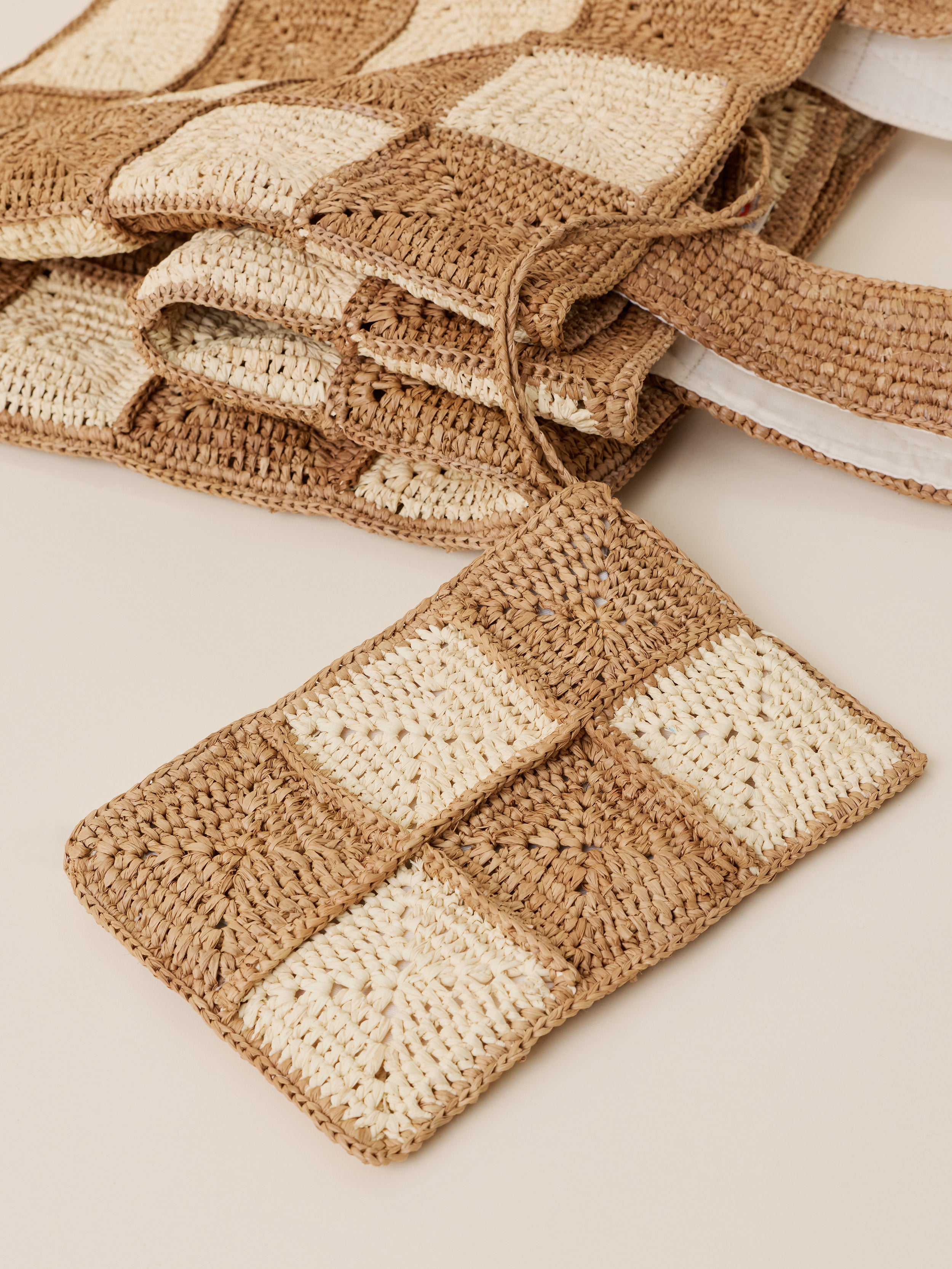 A close-up of a woven checkered pouch or small bag made from tan and cream woven squares, with a matching strap, photographed on a light beige background. More similar woven items are stacked behind it.