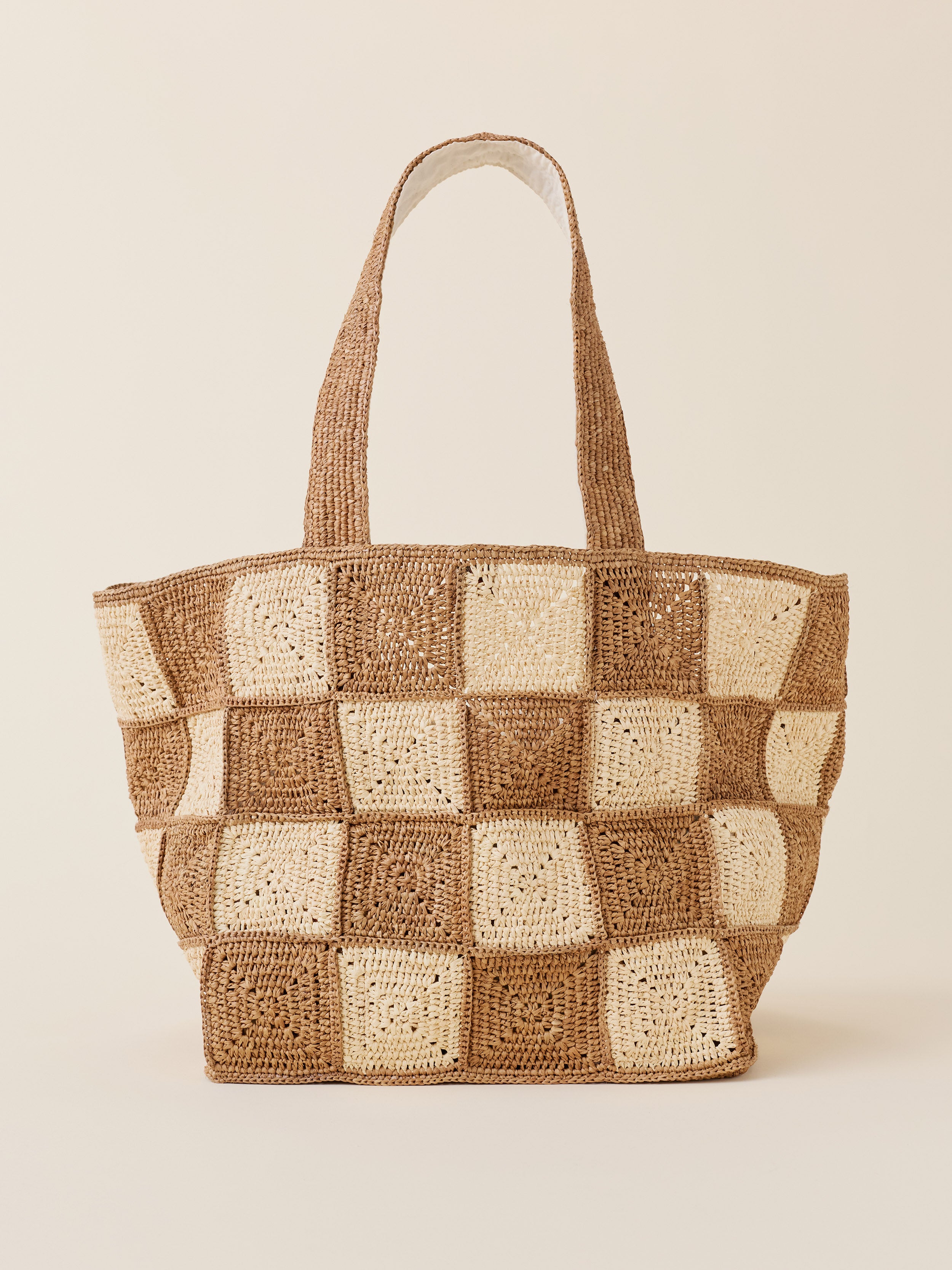 A woven straw tote bag with a checkerboard pattern of natural brown and cream crochet squares, featuring two shoulder straps, set against a solid light beige background.