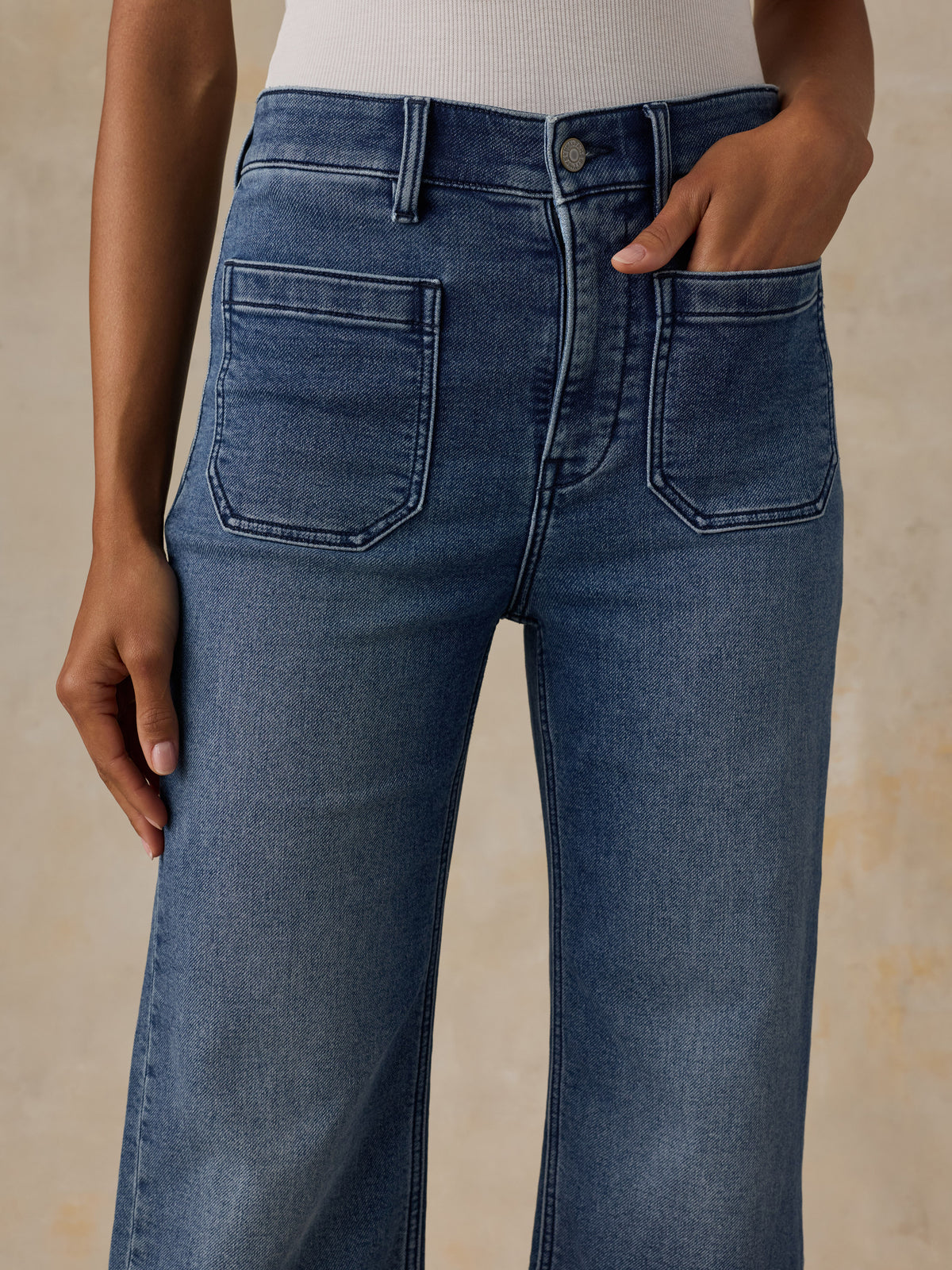 Close-up of a person wearing medium-wash blue denim jeans with a straight-leg or wide-leg fit, shown from the waist down against a warm beige textured background., image 2 of 6