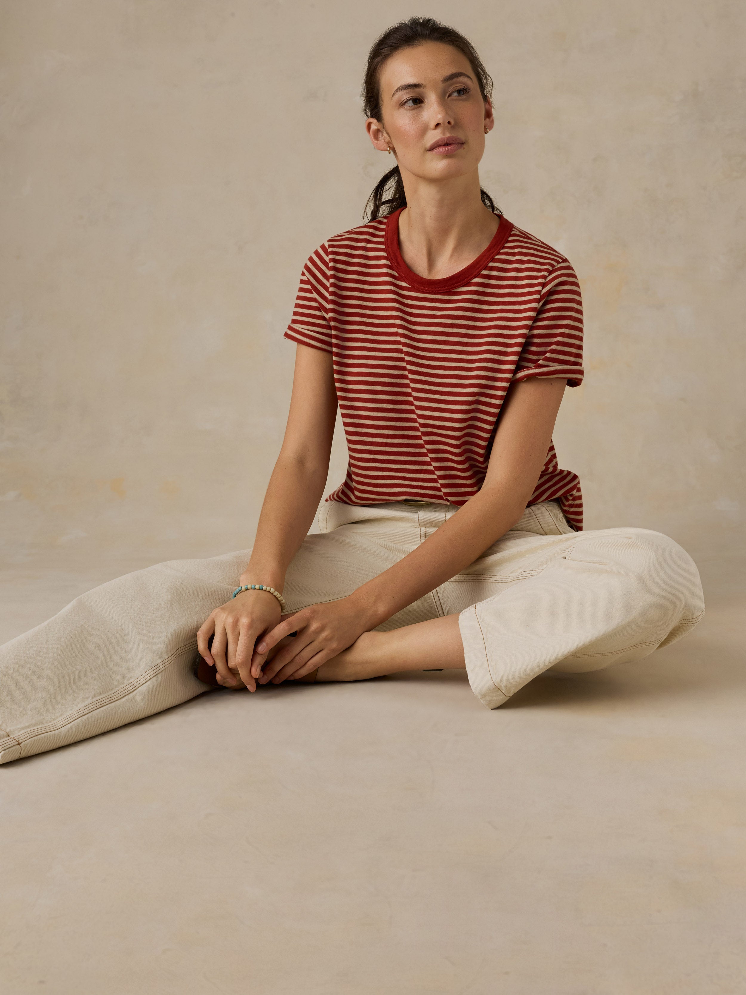 Woman sitting on the ground wearing a red and white striped t-shirt and cream colored denim jeans against a beige background