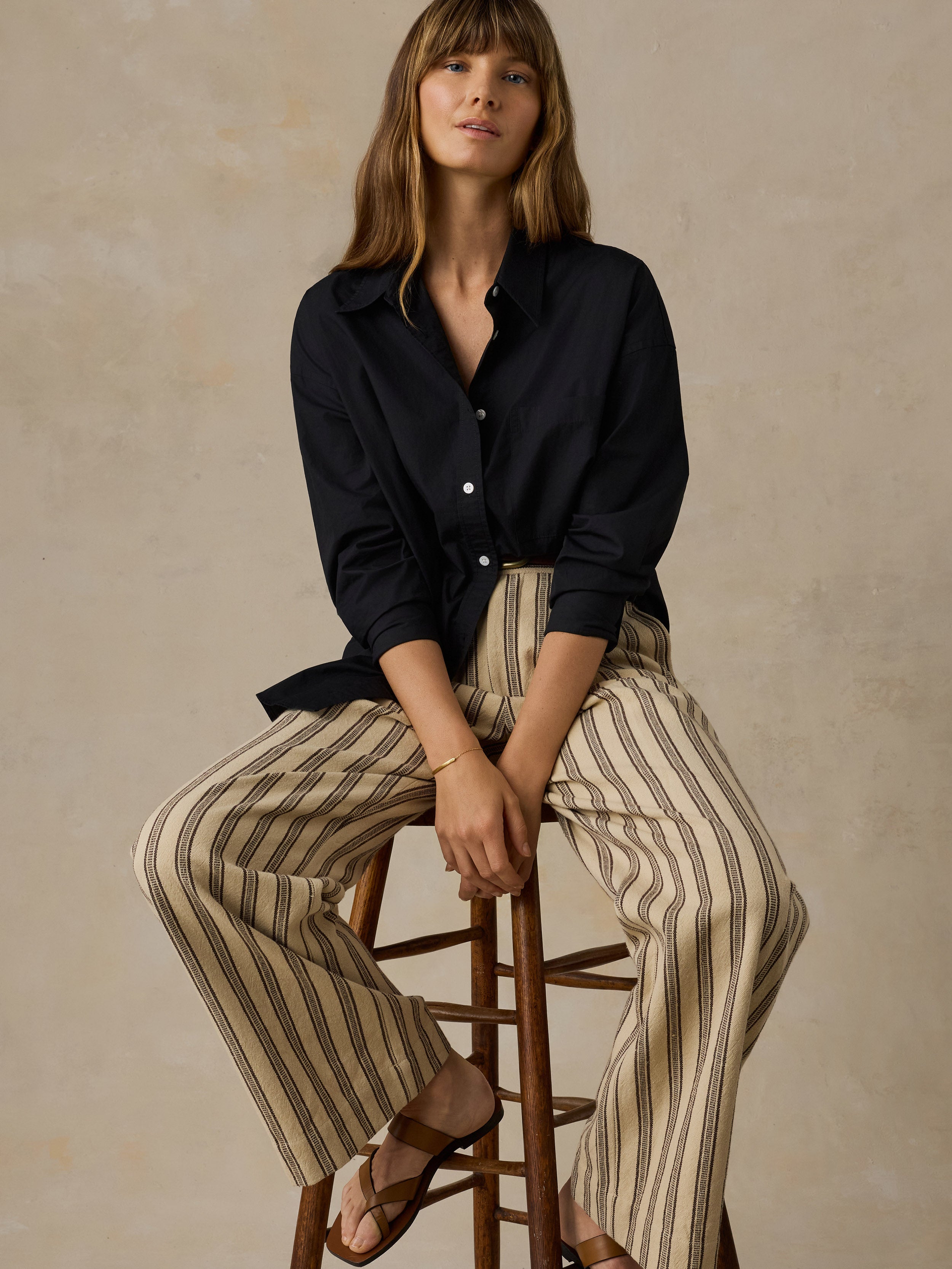 Woman sitting on a wooden stool wearing a black button down shirt and beige and black striped pants against a beige background.