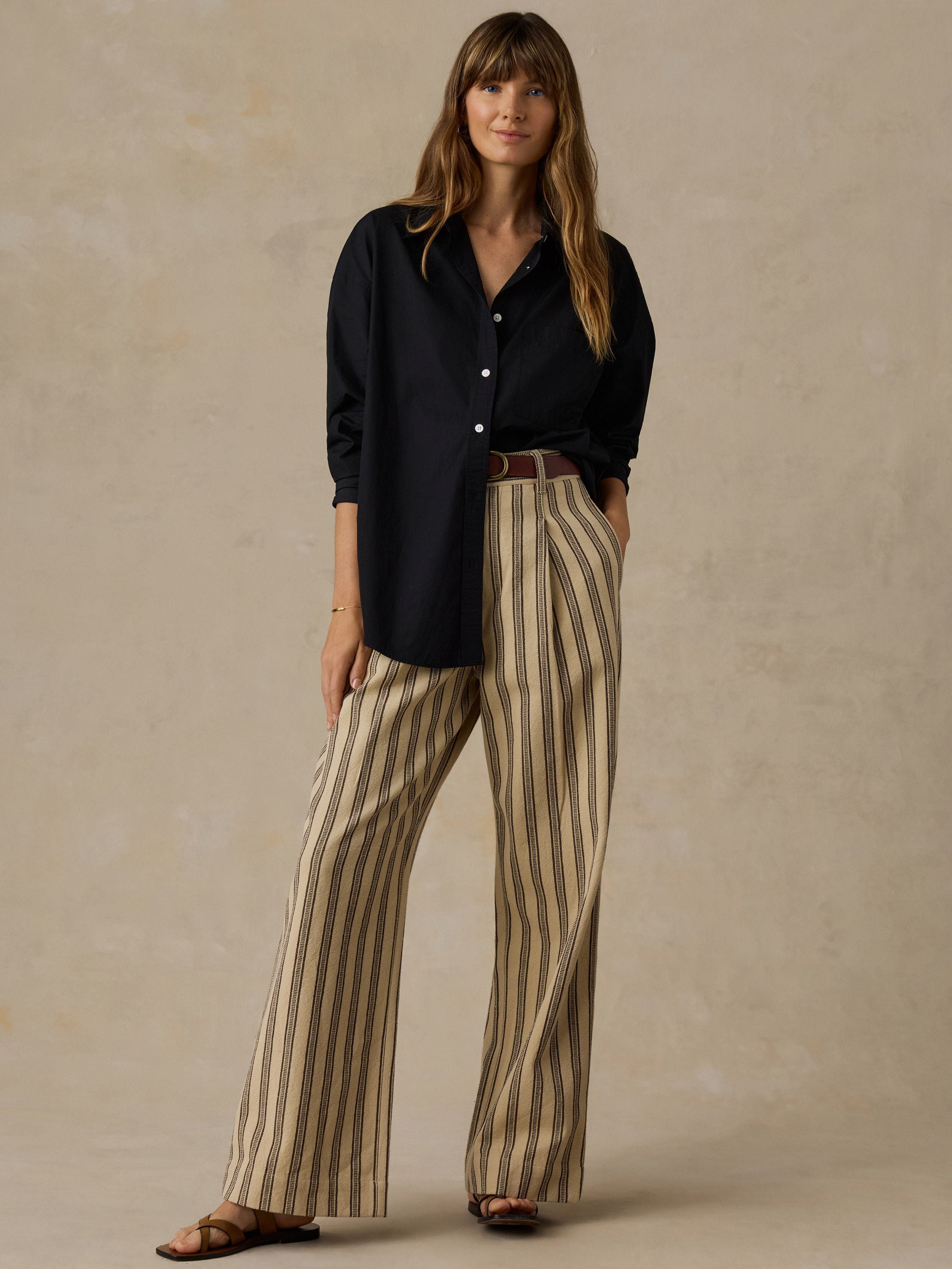 Woman wearing a black button down shirt and beige and black striped pants against a beige background.