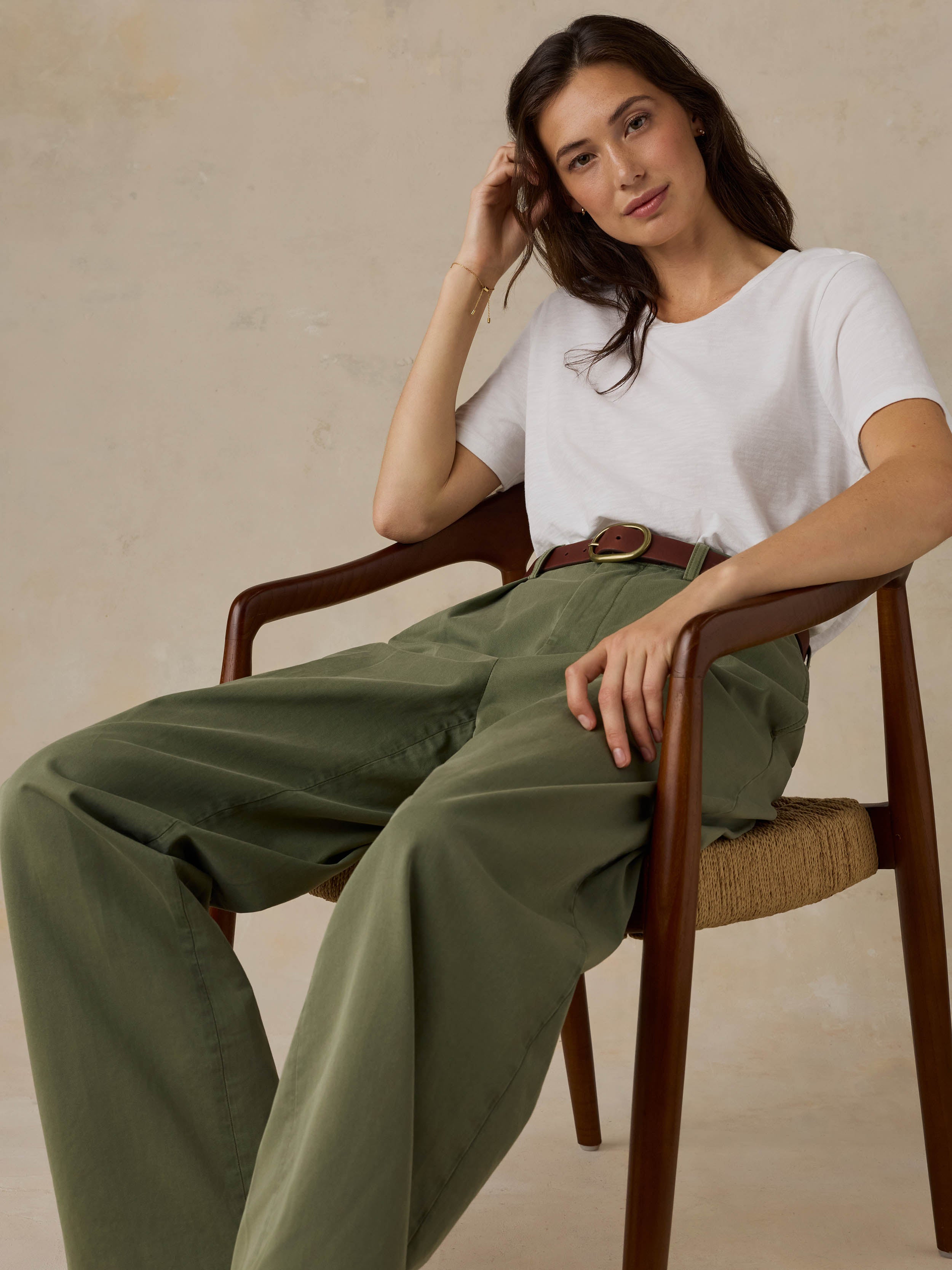 Woman sitting on a wooden chair wearing a white t-shirt and green pants against a beige background