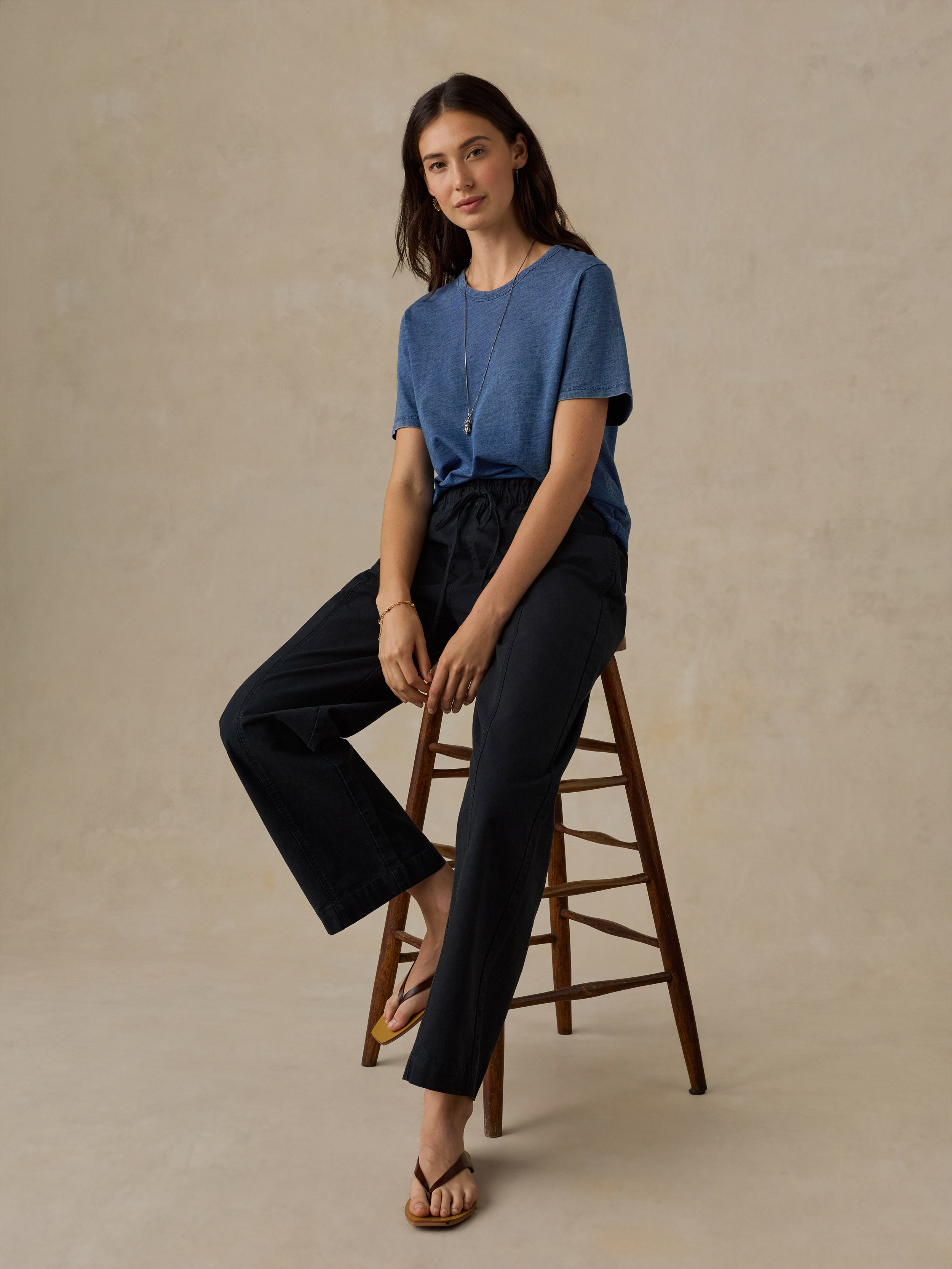 Woman sitting on a wooden stool wearing a blue t-shirt and black drawstring pants against a beige background