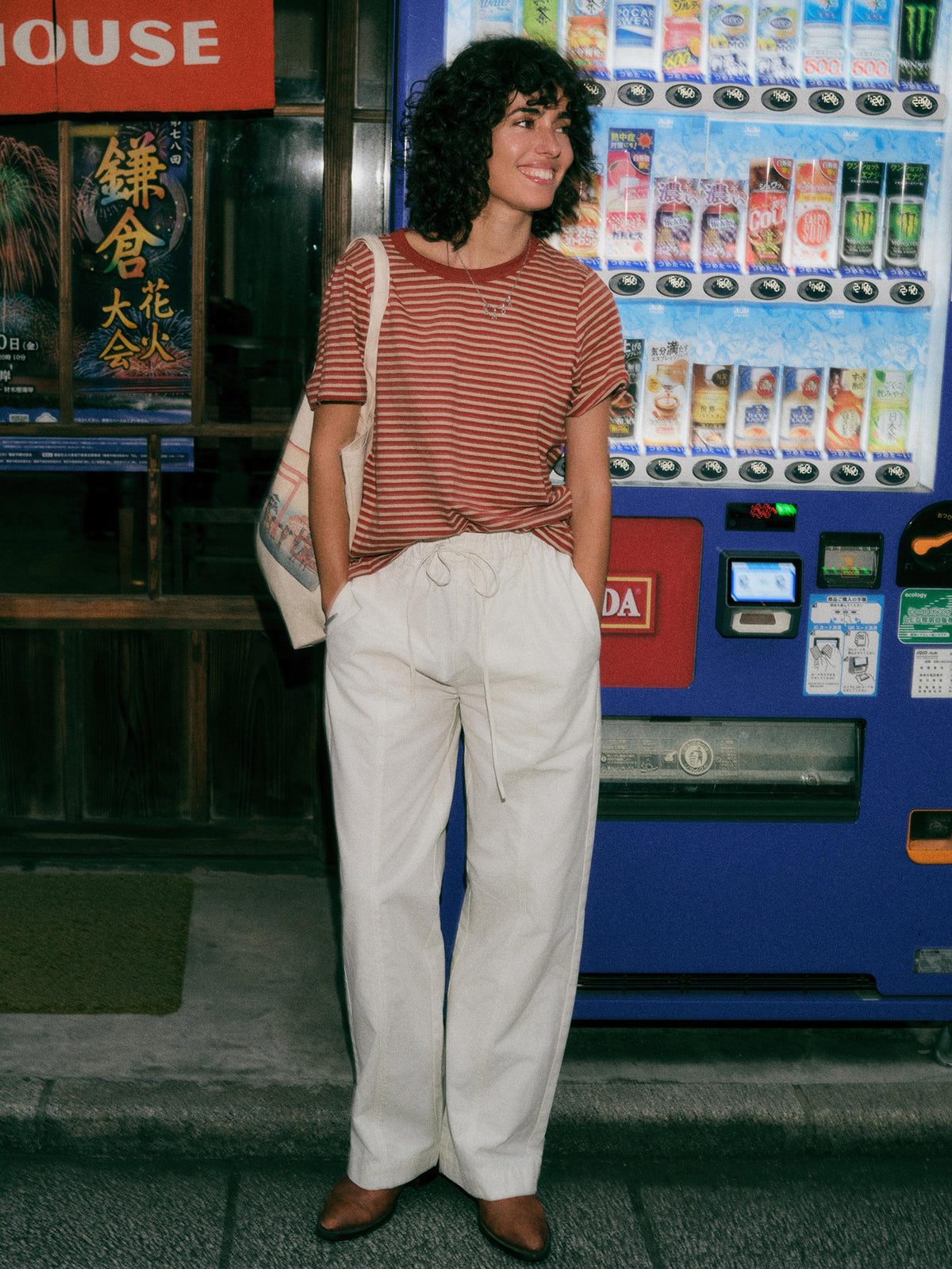 A woman stands in front of a vending machine wearing a rust-red and white striped t-shirt, cream wide-leg drawstring pants, and brown shoes, with a canvas tote bag slung over her shoulder., image 1 of 6