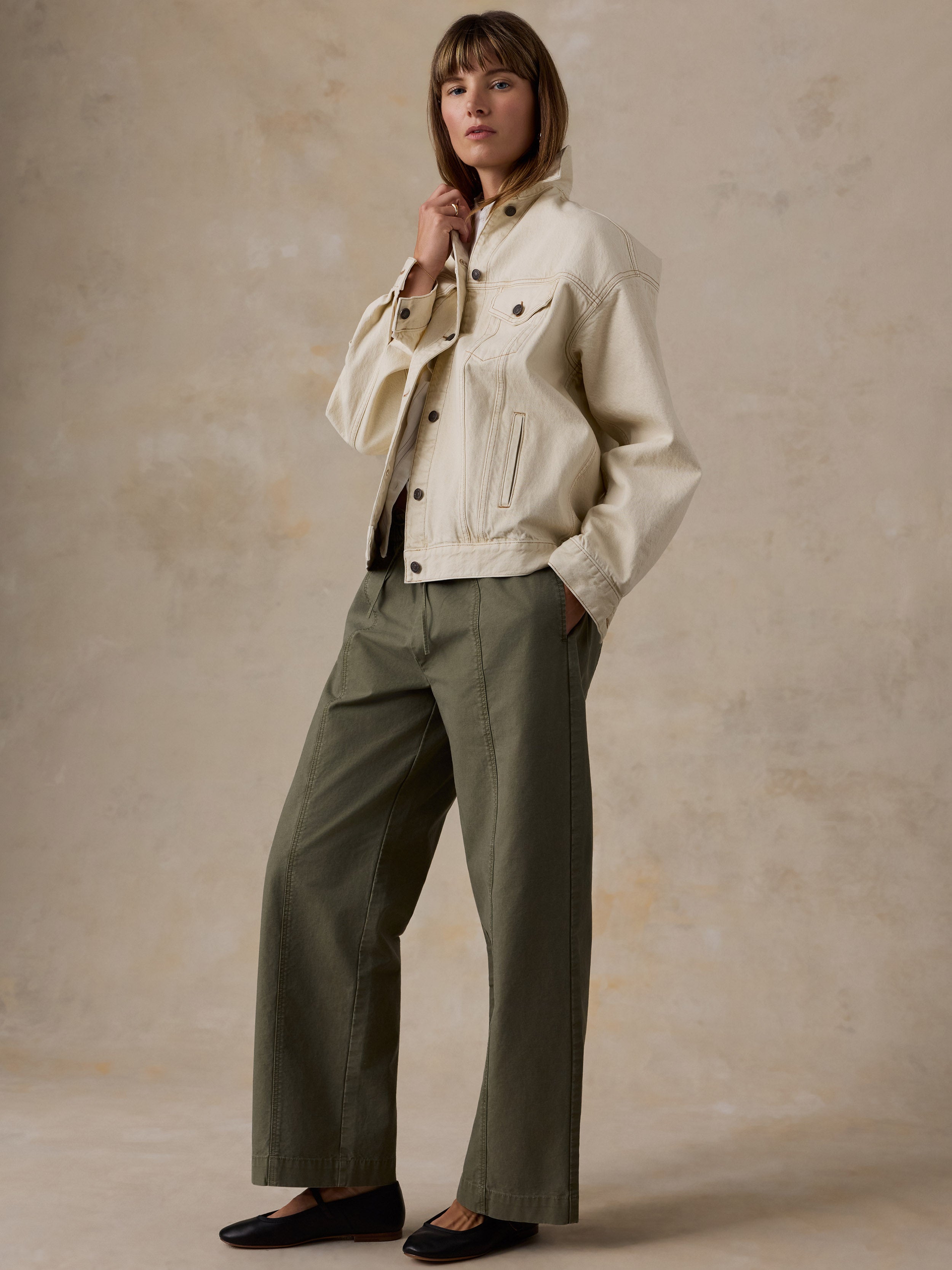 A woman wearing an off-white denim jacket and olive green pants against a beige studio background