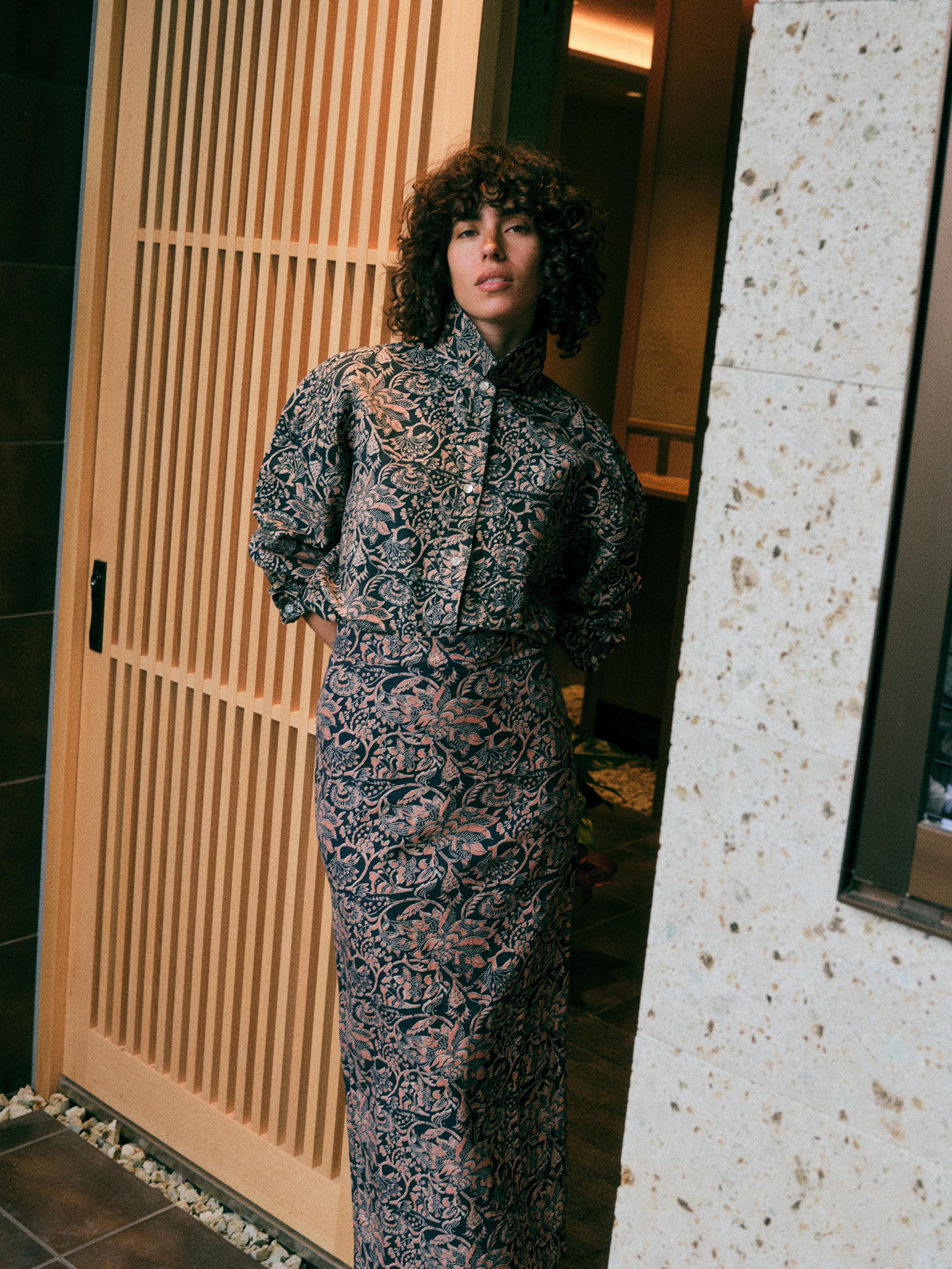 A woman with curly dark hair stands in a doorway wearing a long-sleeved, high-neck midi dress with a dense abstract floral pattern in black, navy, and dusty pink. The background features a light wood slatted door on the left and a textured stone pillar on the right.