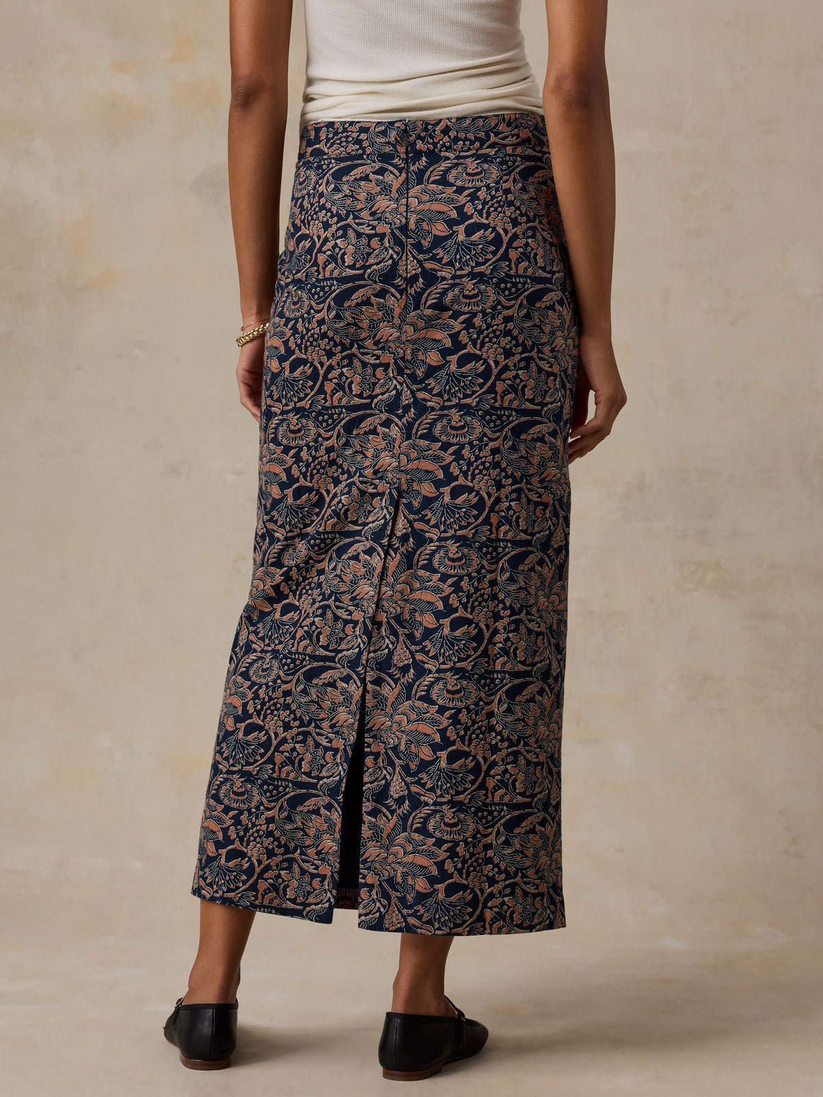A model is shown from the back wearing a dark navy blue maxi skirt with a beige floral pattern, paired with a cream top and black shoes, set against a textured beige background., image 4 of 6
