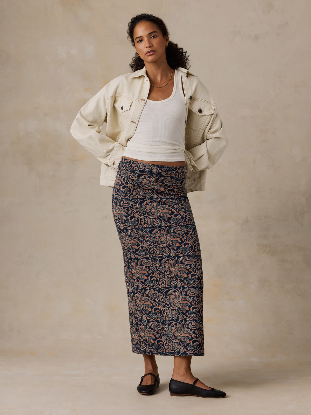A woman stands against a textured beige studio wall wearing a cream jacket over a white ribbed tank top and a long navy skirt with a tan floral pattern. She is wearing black flats., image 6 of 6