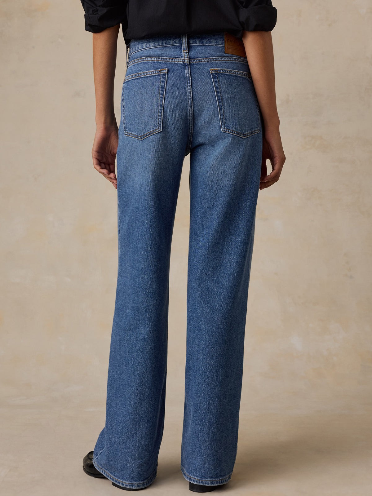 A woman shown from the back wearing blue wide-leg jeans and a white blouse, standing against a warm beige textured studio backdrop., image 2 of 6
