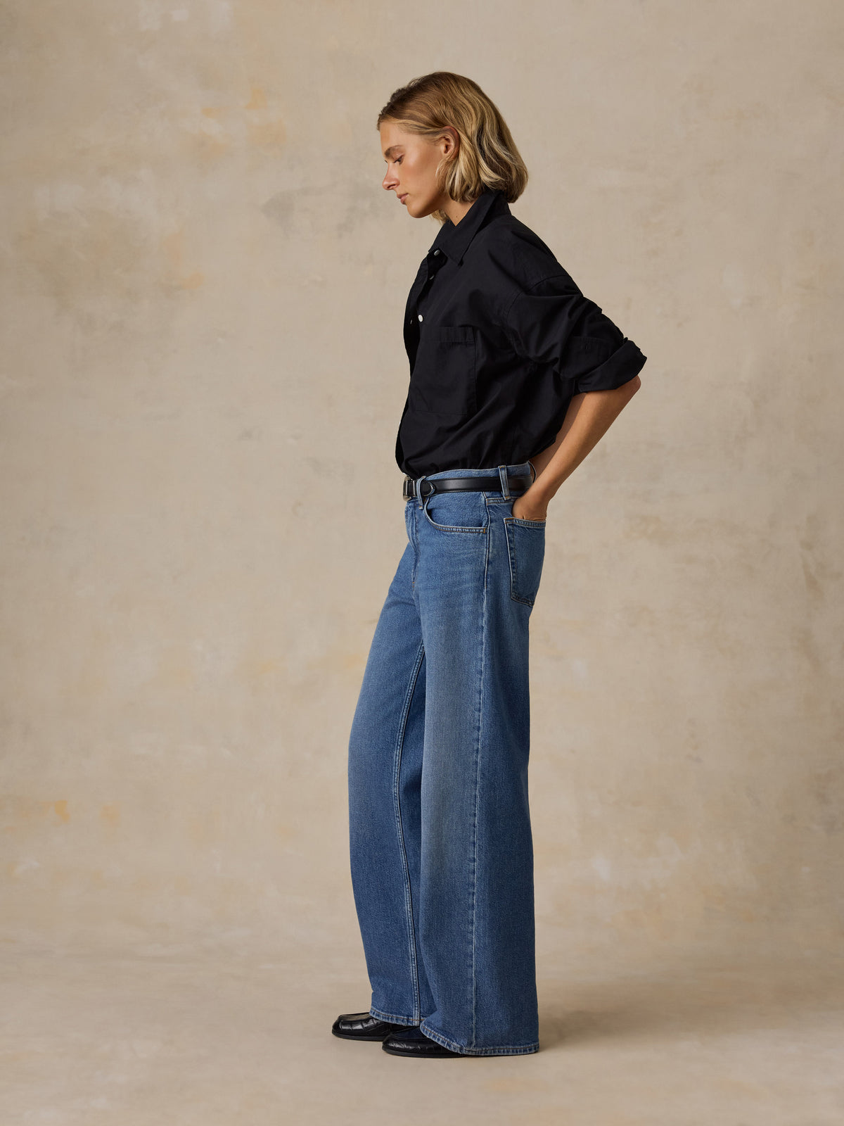 A woman sits on a beige floor against a textured beige wall, wearing a white button-up shirt, blue wide-leg jeans, and black Mary Jane flats., image 5 of 6