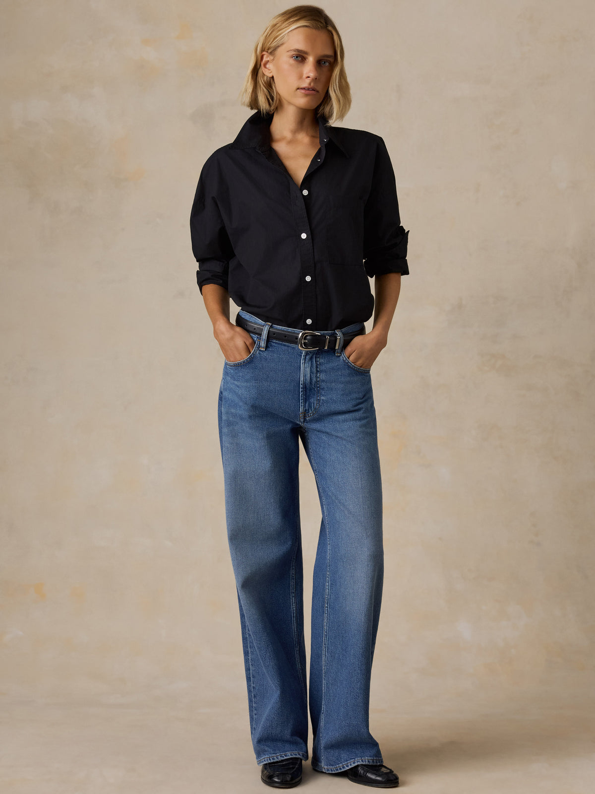 A woman stands against a textured beige studio backdrop wearing a white button-up shirt with rolled sleeves, blue straight-leg jeans, and black flats., image 6 of 6