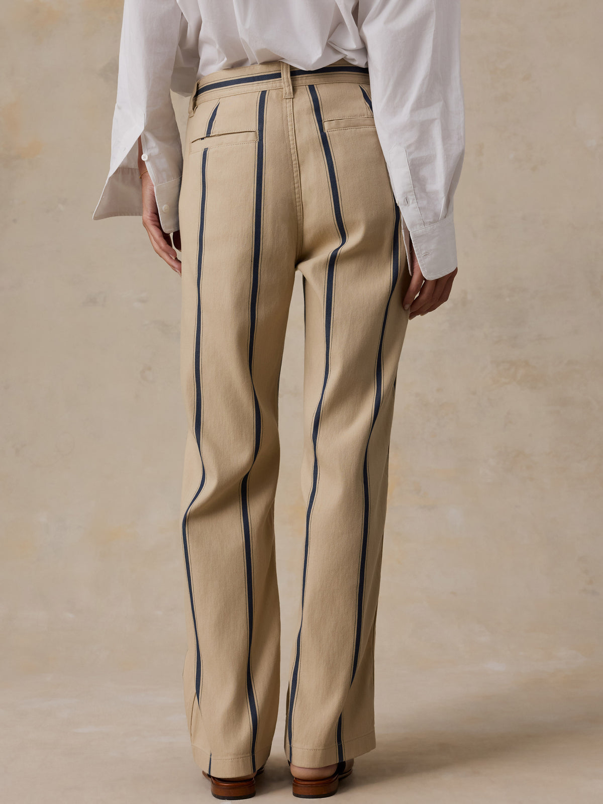 A person shown from the back wearing beige trousers with dark vertical pinstripes, a white long-sleeve shirt, and brown shoes against a warm beige textured studio background., image 4 of 8
