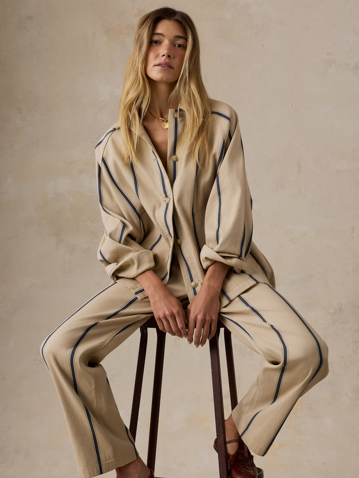 A woman sits on a wooden stool wearing a beige matching lounge set with thin dark blue vertical stripes. The set includes a long-sleeve button-front top and wide-leg pants, photographed against a textured beige studio backdrop., image 3 of 8