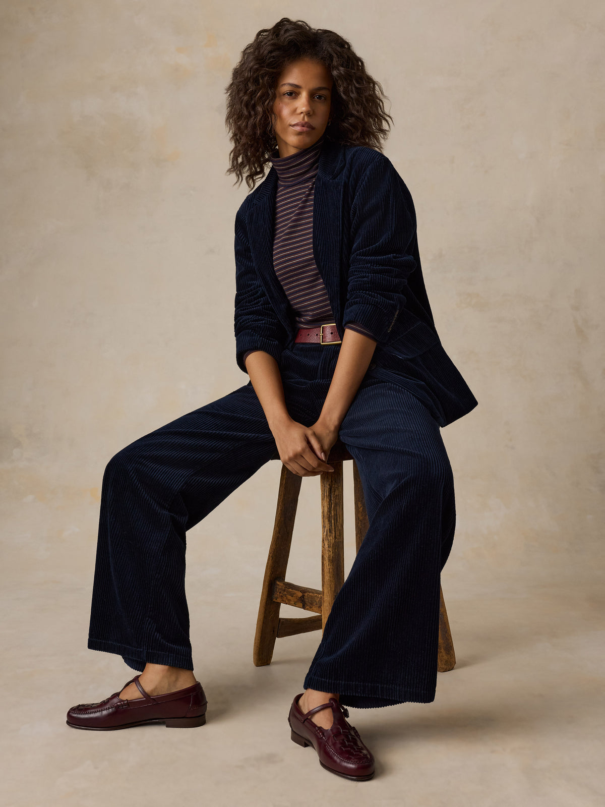 A woman sitting on a stool wearing a dark navy corduroy suit and a striped turtleneck, image 3 of 7