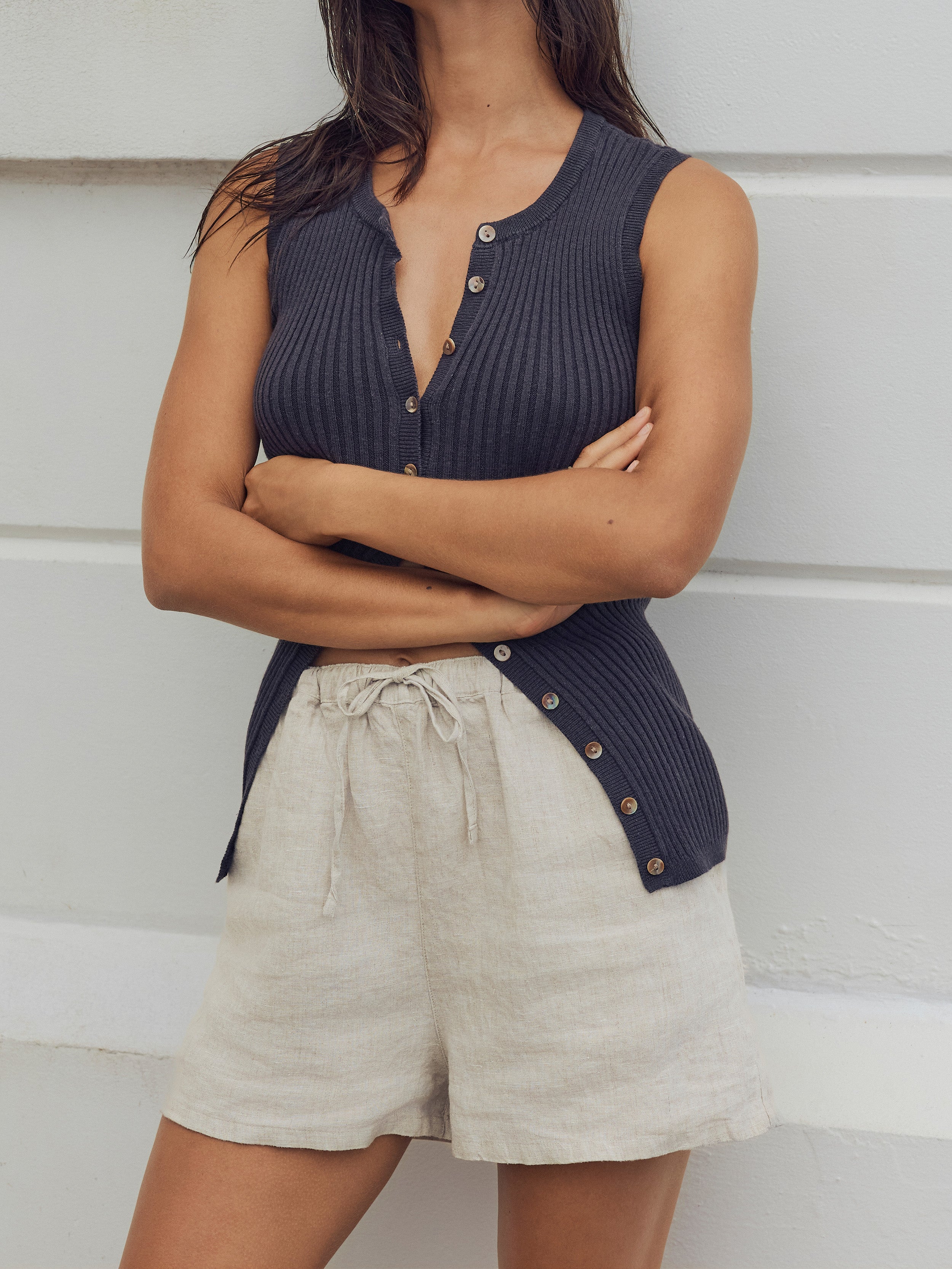 Woman wearing beige linen shorts and a black tank top standing against a white wall