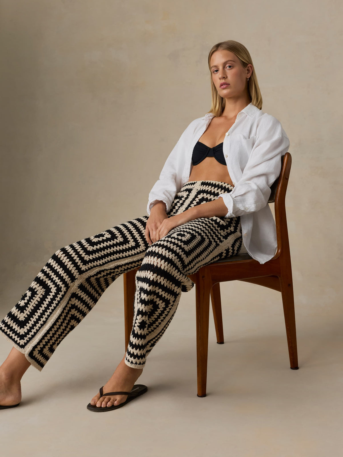 Woman sitting on a chair wearing a white cardigan, black and white patterned pants, and a black bralette., image 3 of 5