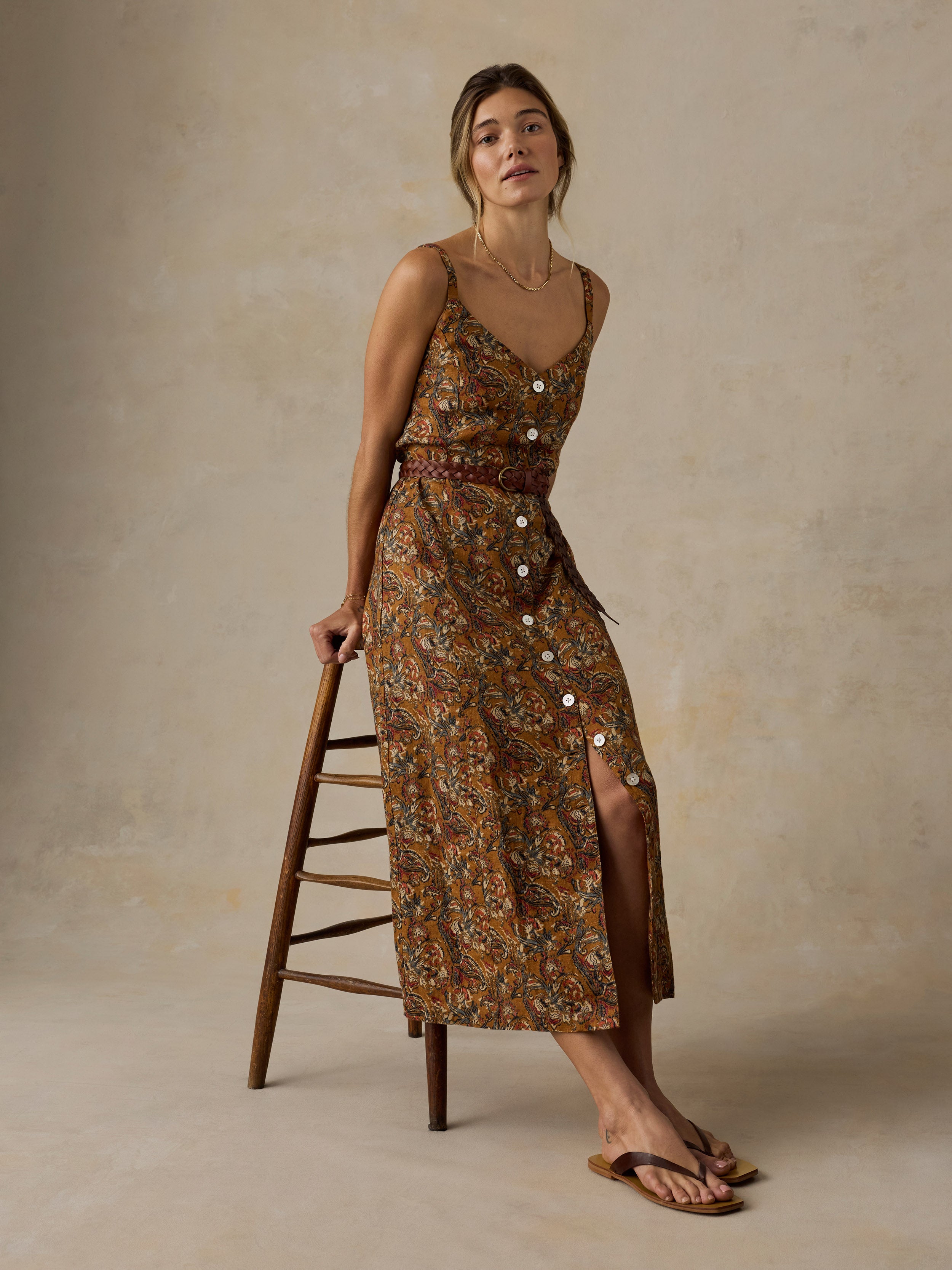 Woman sitting on a wooden stool wearing an orange and brown printed dress and sandals against a beige background