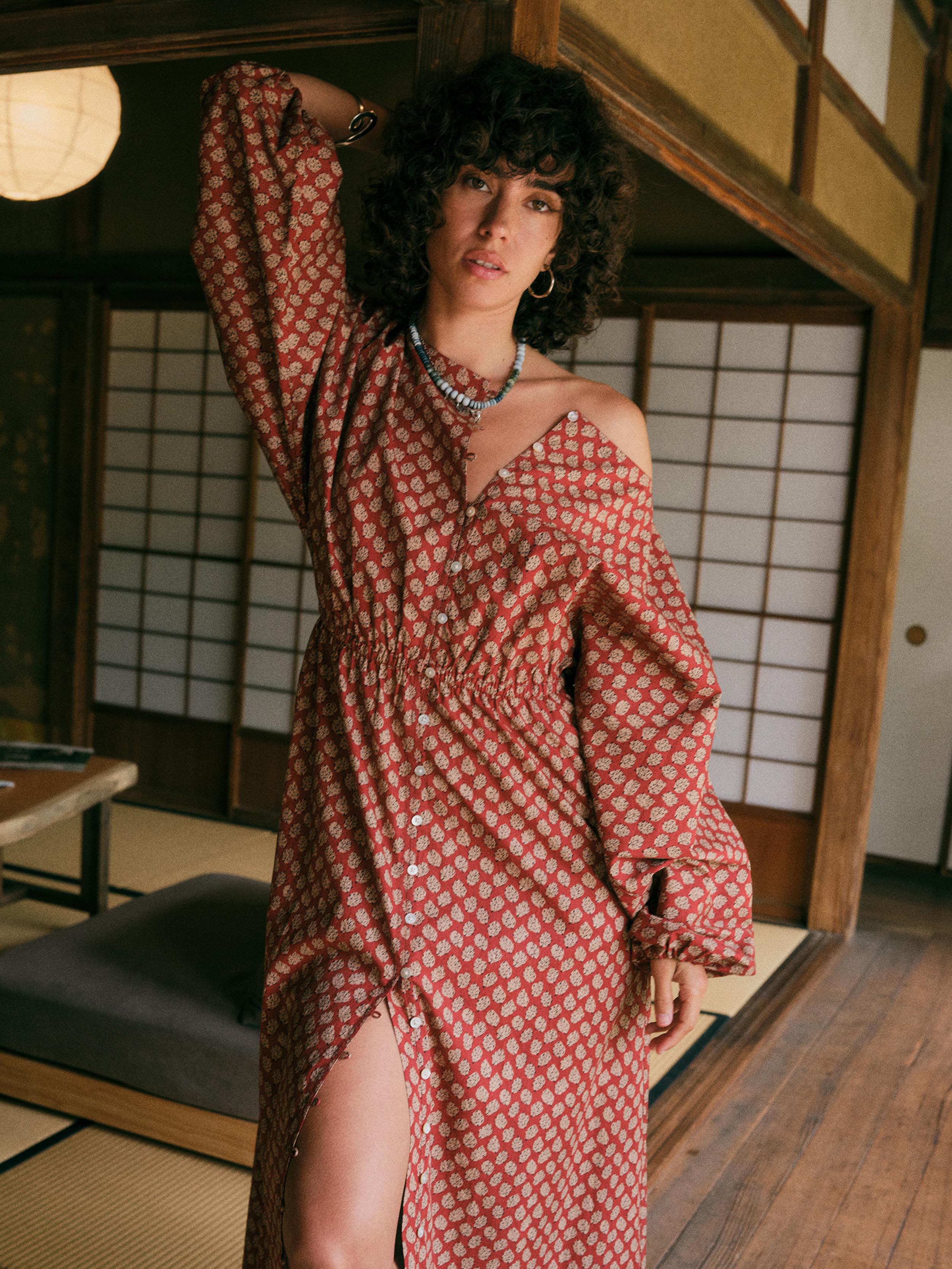 A woman with curly dark hair models a rust-red off-the-shoulder maxi dress with a white floral pattern, set in a traditional Japanese room with shoji screens.