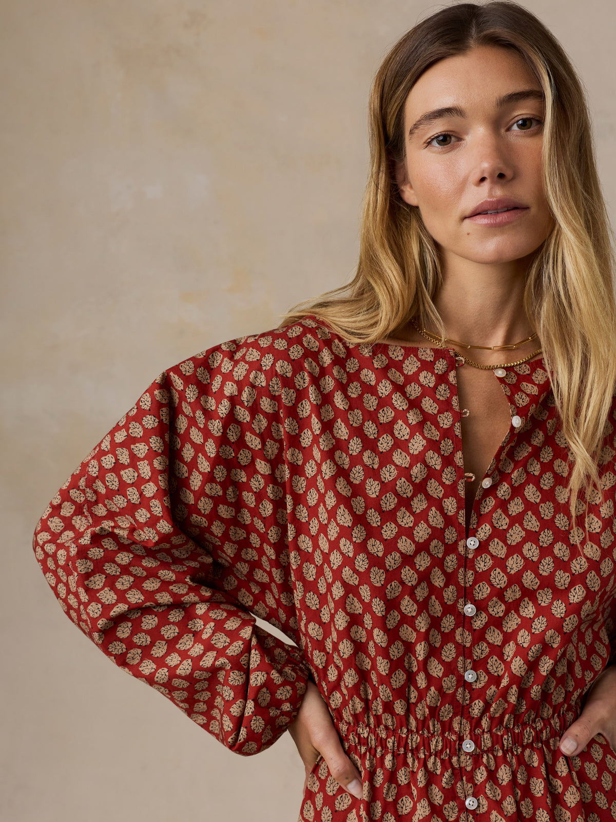 A woman wearing a red long-sleeve button-up dress with a beige floral block print, photographed against a neutral textured beige background., image 6 of 6