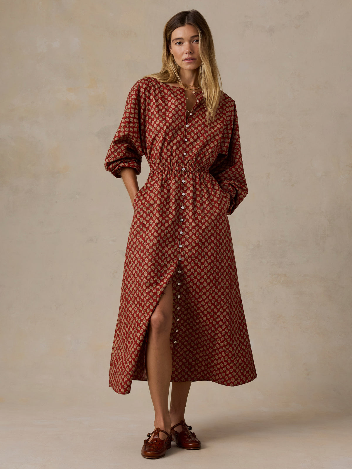 A woman modeling a rust-red midi shirt dress with a dark geometric pattern, button-down front, and a high side slit, paired with brown leather shoes against a textured beige studio backdrop., image 2 of 6